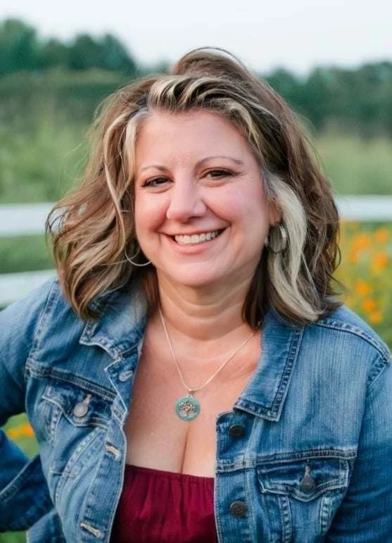 A smiling woman with curly, shoulder-length hair wearing a denim jacket and a red top, outdoors with a blurred green landscape in the background.