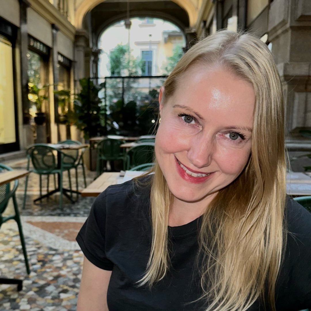 A woman with blonde hair and blue eyes smiling while sitting in a cafe with outdoor seating, decorated with chairs and tables, in a city with historic architecture.
