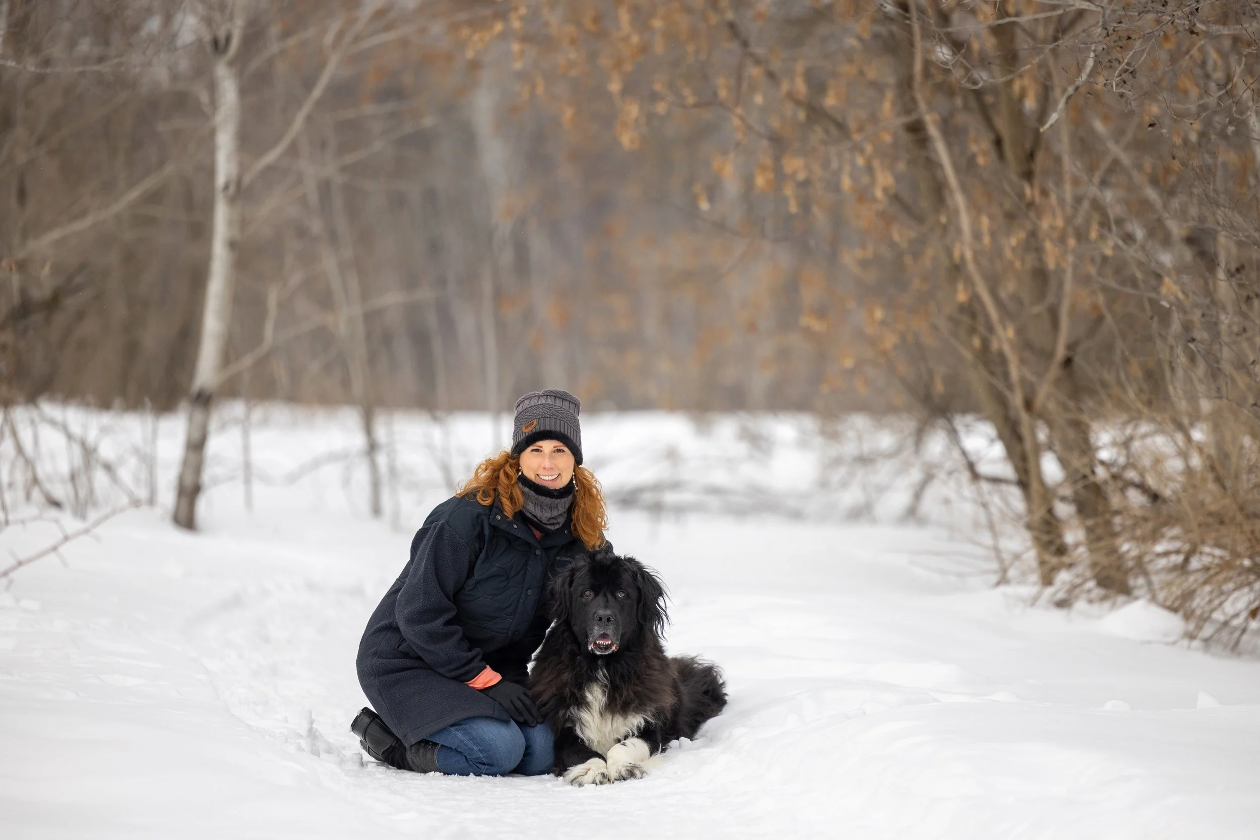 A woman with red hair wearing a gray beanie, black coat, and gloves kneeling on snow-covered ground next to a large black and white dog, in a winter forest with leafless trees.