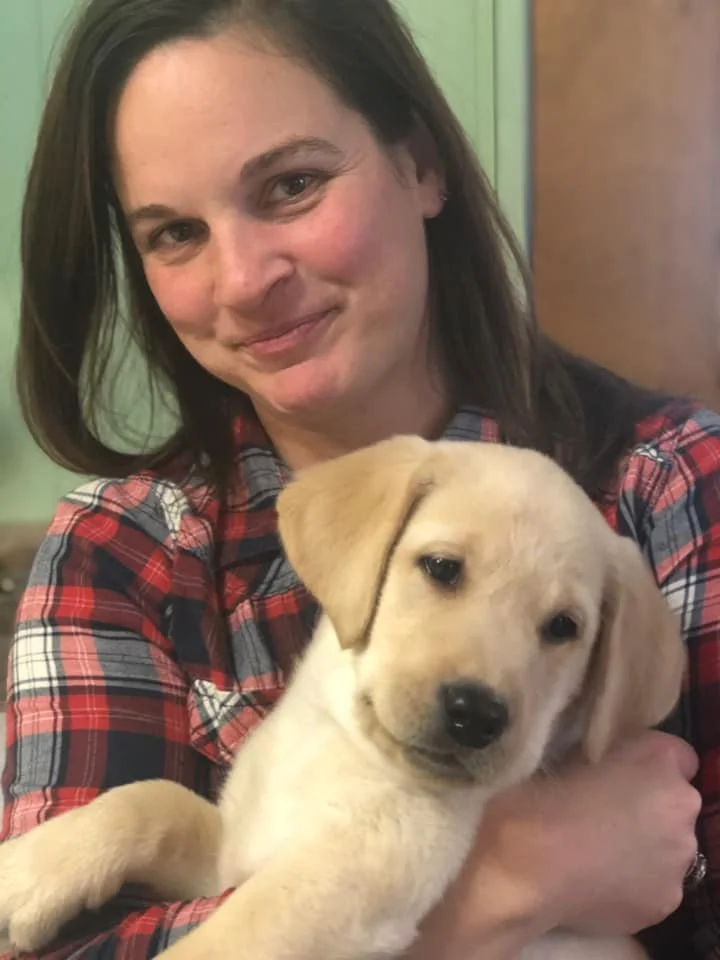 A woman with dark hair holding a yellow Labrador Retriever puppy indoors.