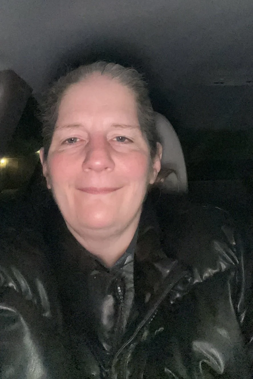 A woman with light skin, tied-back brown hair, smiling, wearing a black leather jacket, sitting inside a car at night.