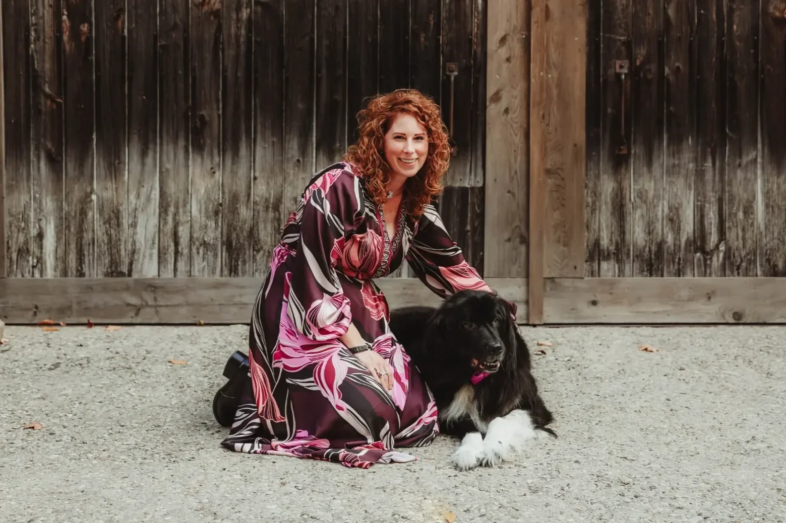 A woman with curly red hair in a floral dress kneeling on the ground beside a black and white dog in front of a wooden fence.