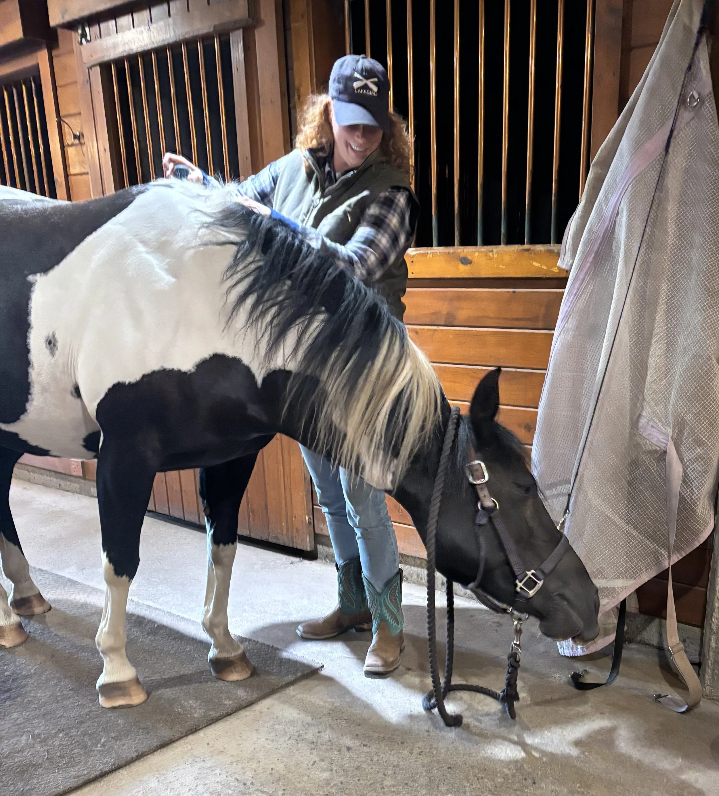 A woman in western attire, including a cap, vest, and cowboy boots, is grooming a black and white horse inside a wooden stable.
