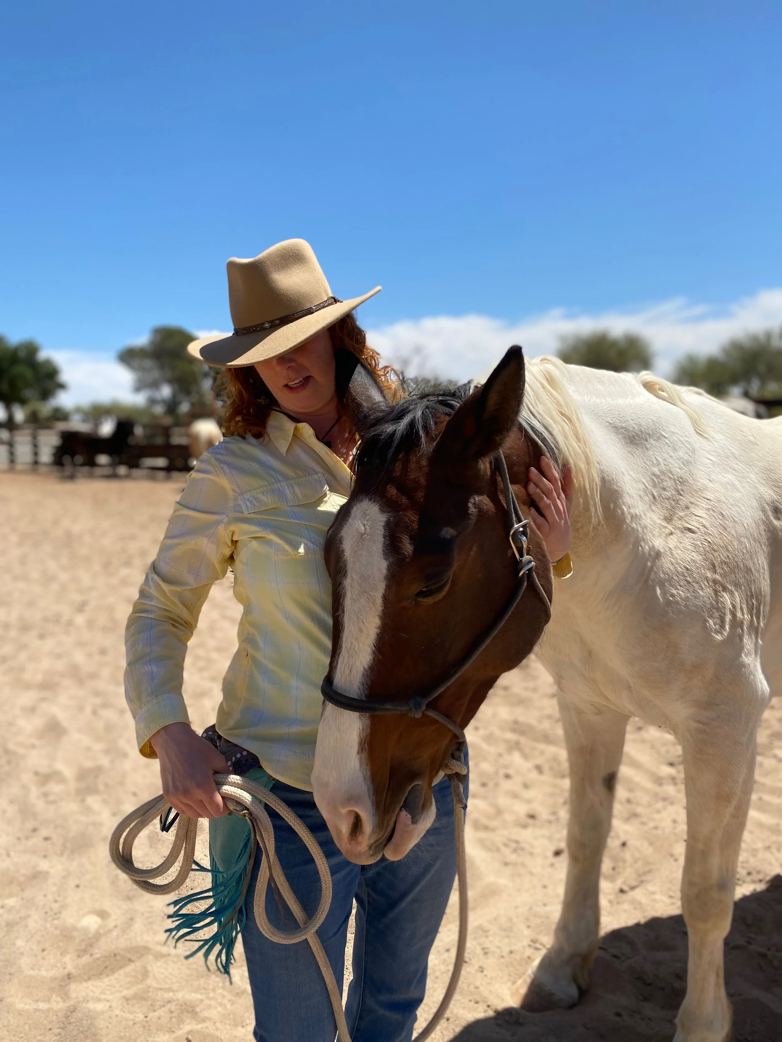 A woman wearing a beige hat, a yellow shirt, and jeans standing on a sandy area, holding a brown and white horse by the reins and gently petting its head.