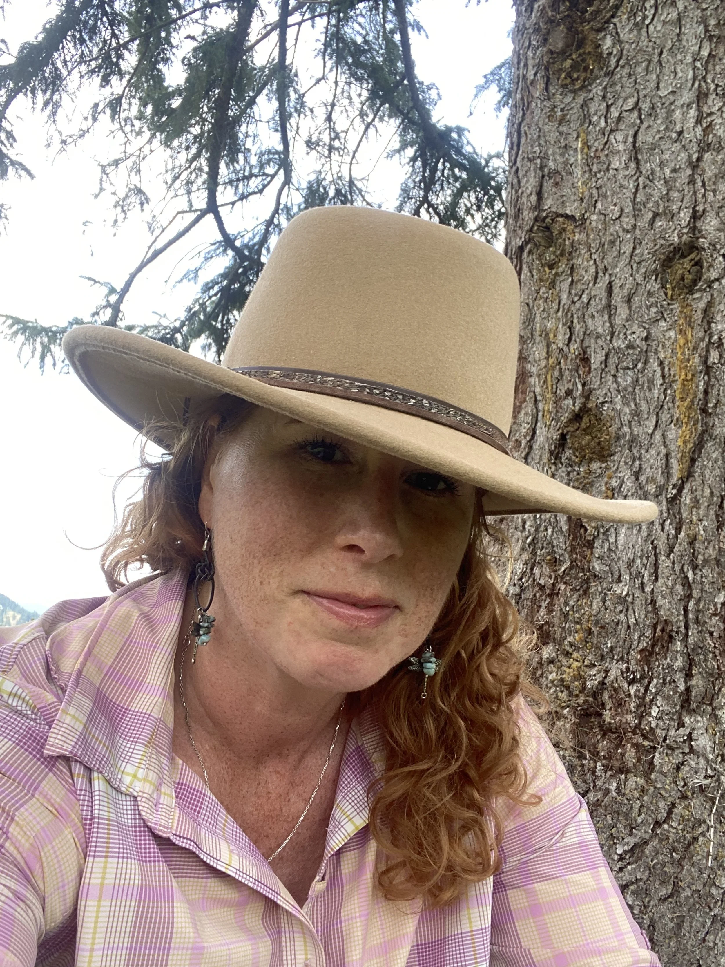 A woman with curly red hair wearing a wide-brimmed beige hat with a leather band, earrings, and a light-colored plaid shirt standing outdoors next to a tree with a textured bark.