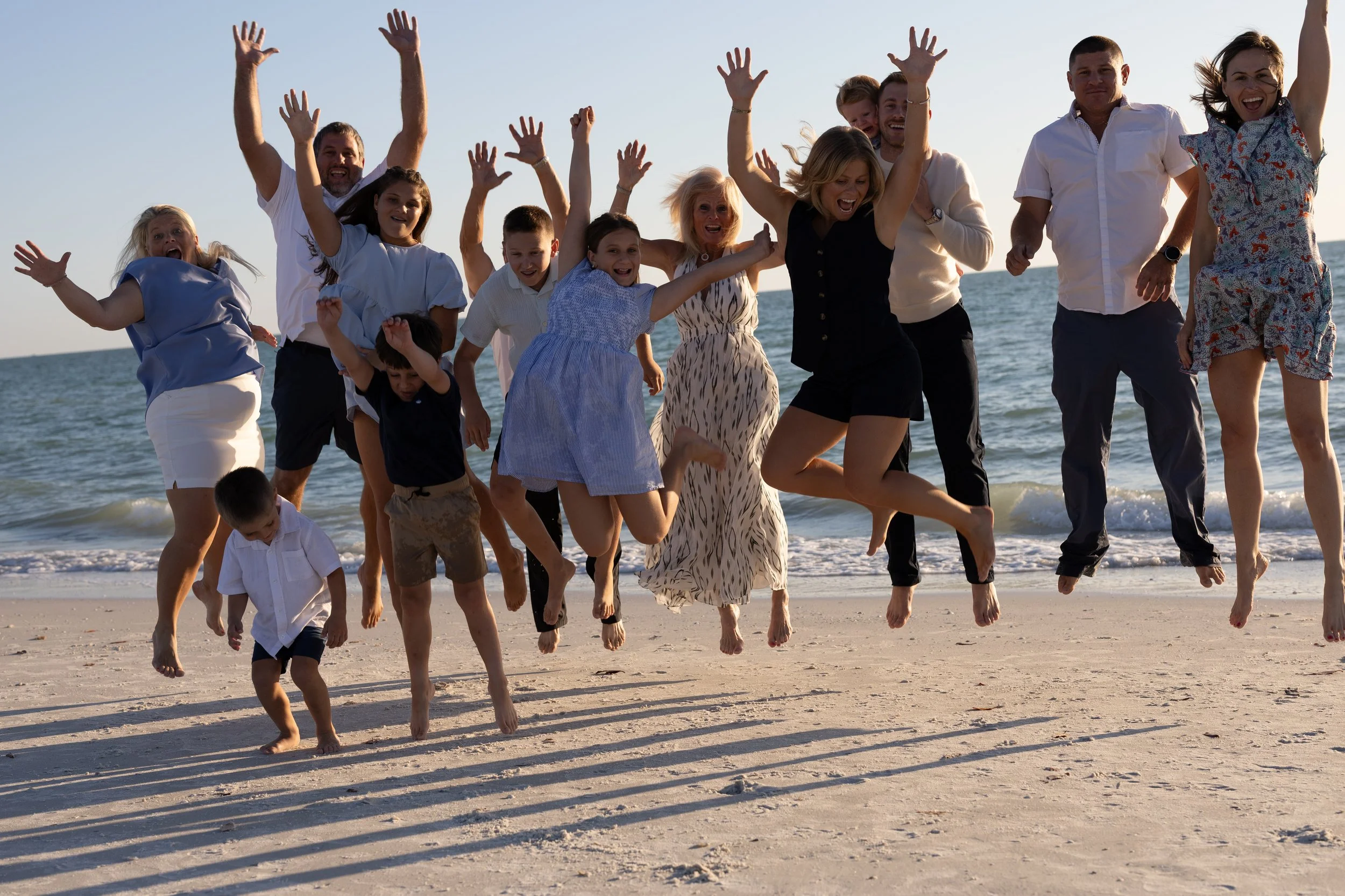 A large group of people, including children and adults, are jumping and celebrating on a sandy beach near the ocean during sunset or late afternoon.
