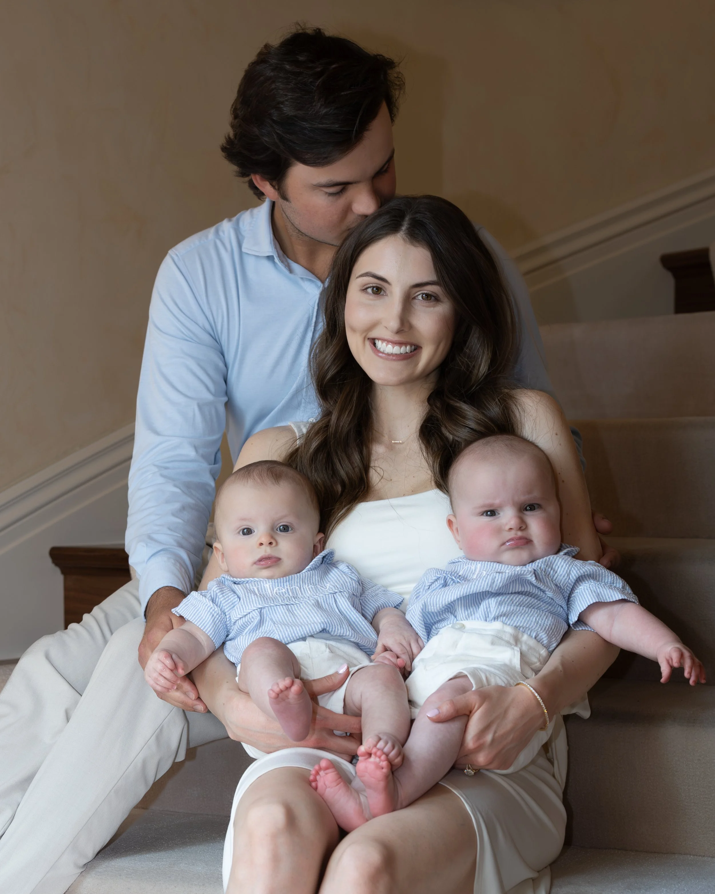 A woman sitting on the floor with two babies on her lap, a man standing behind her kissing her on the forehead.