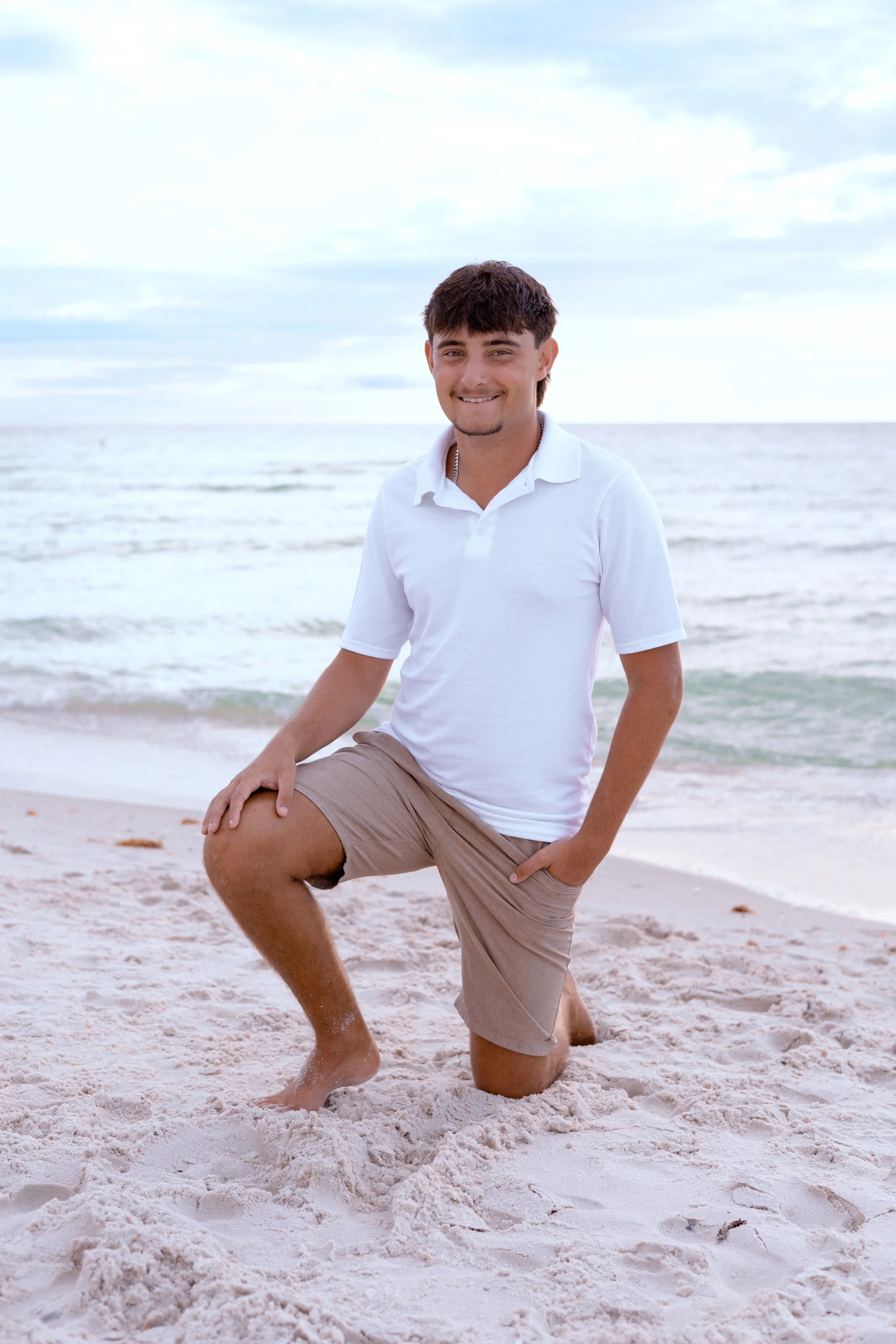 A young man kneeling on the beach with one knee in the sand, smiling, wearing a white polo shirt and tan shorts, with the ocean and sky in the background.