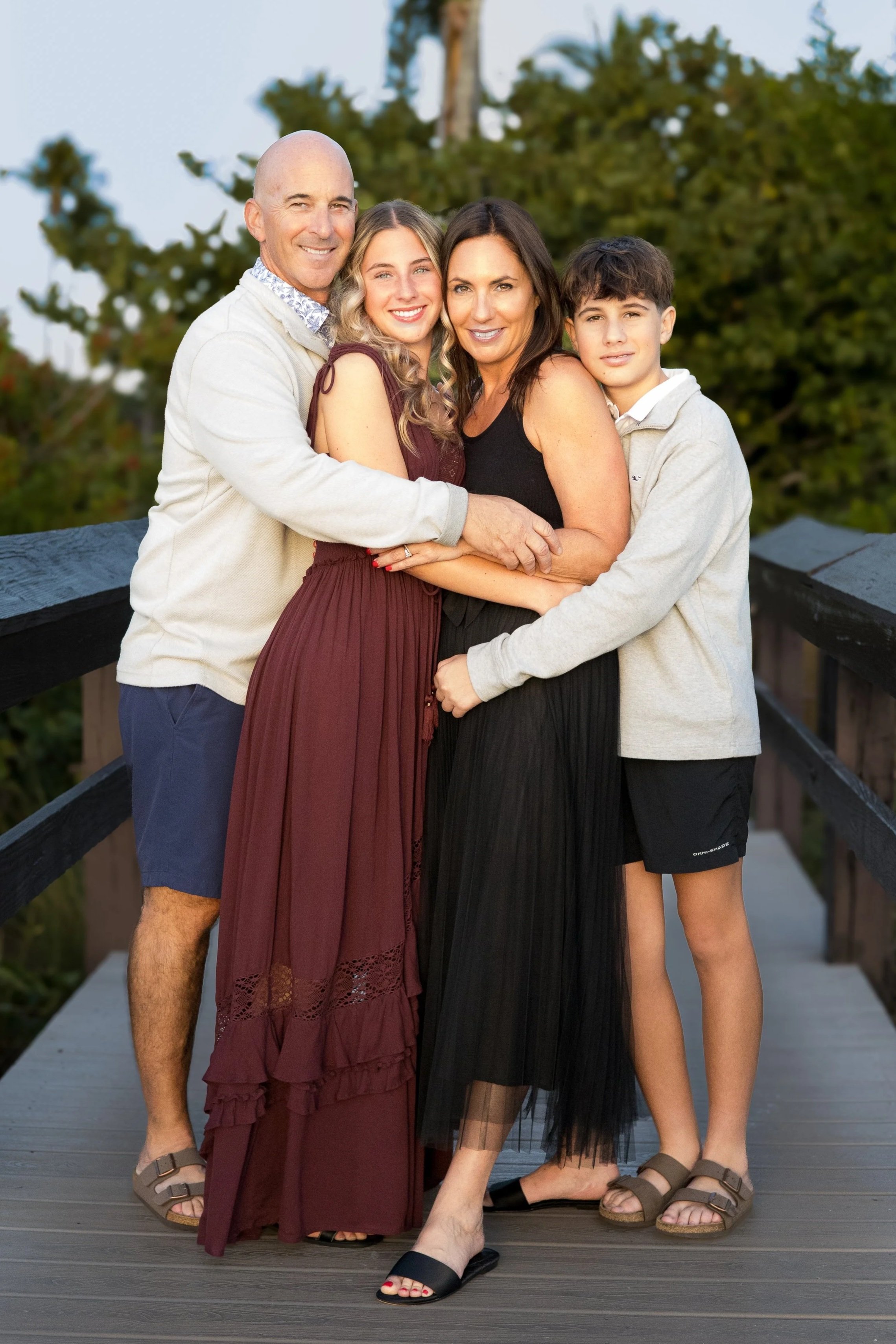 A family of four standing on a wooden bridge outdoors, smiling and hugging. The family includes a senior man, a young woman, a middle-aged woman, and a teenage boy, with trees in the background.