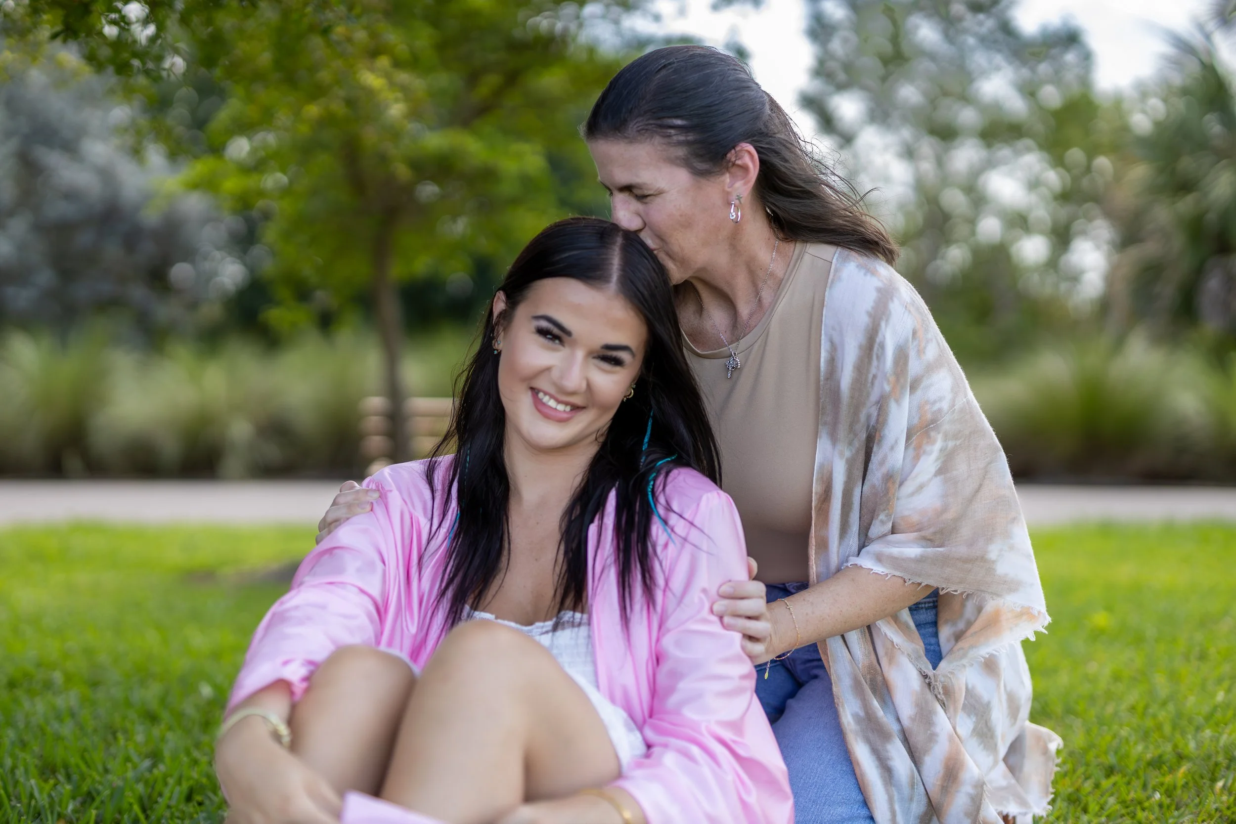 A young woman in a pink jacket sitting on grass, smiling, with an older woman standing behind her, kissing her forehead, in a park with trees and green grass.
