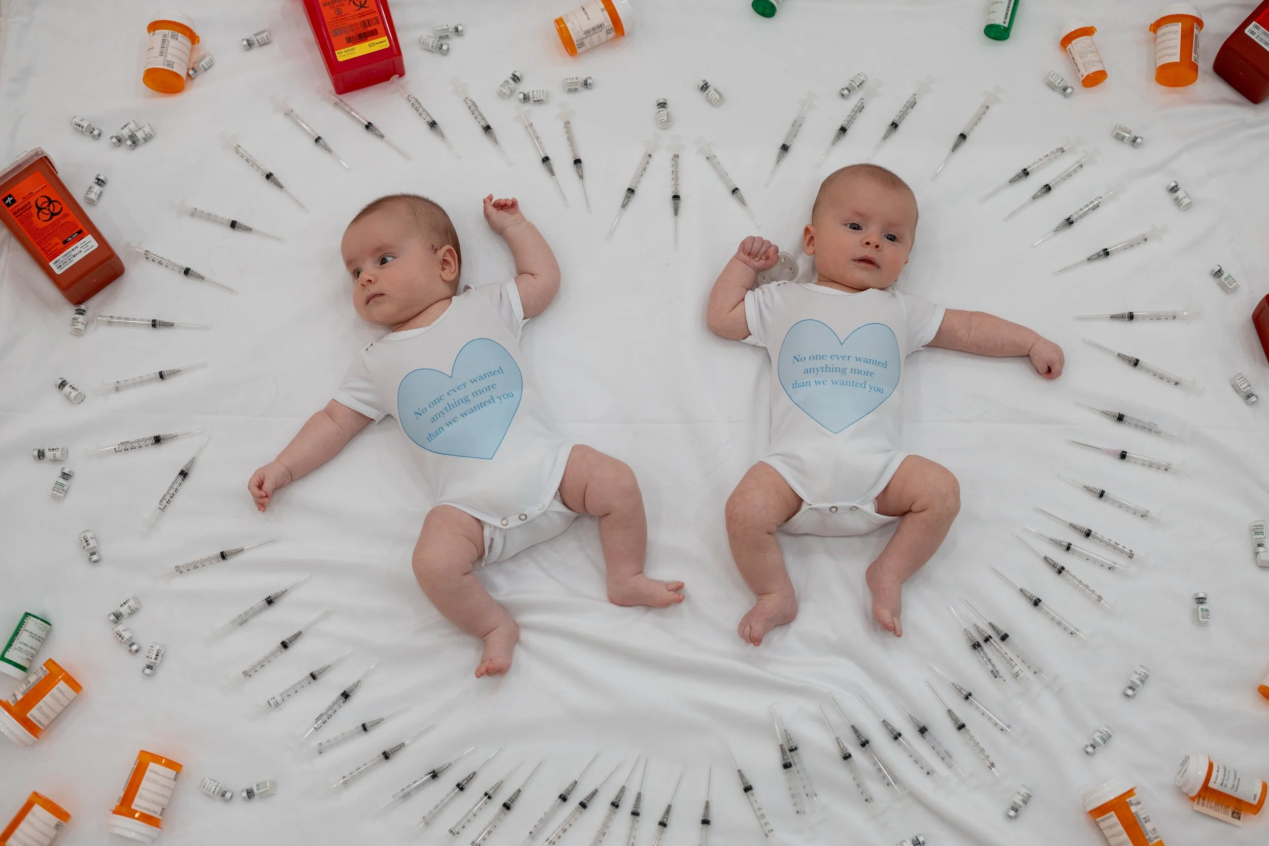 infertilty success picture of Two babies lying on a white surface, surrounded by medicine bottles, syringes, and medical supplies. They are wearing white onesies with blue heart-shaped labels that contain text.