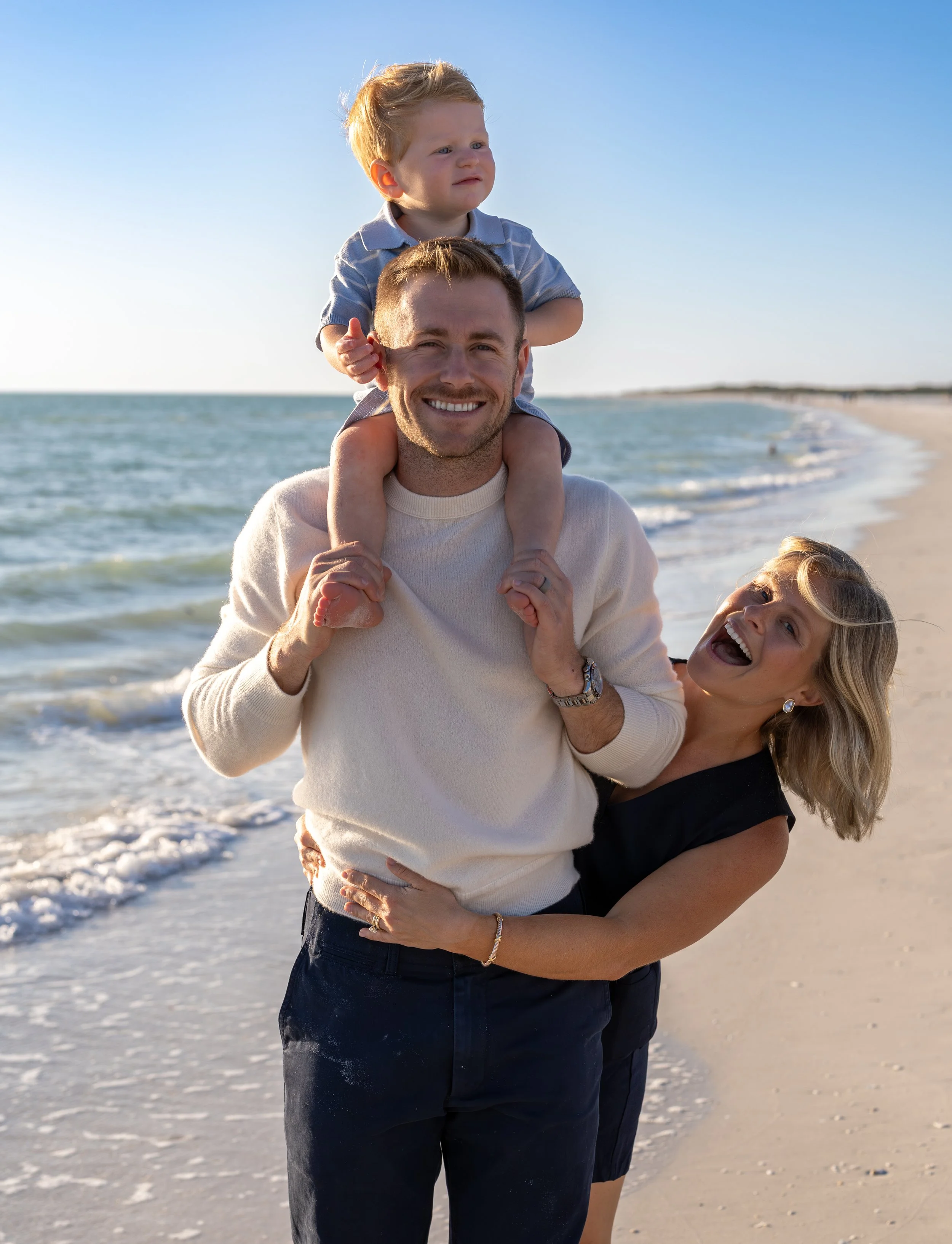 A happy man carrying a young boy on his shoulders at the beach, with a woman standing nearby, all enjoying a sunny day by the ocean.