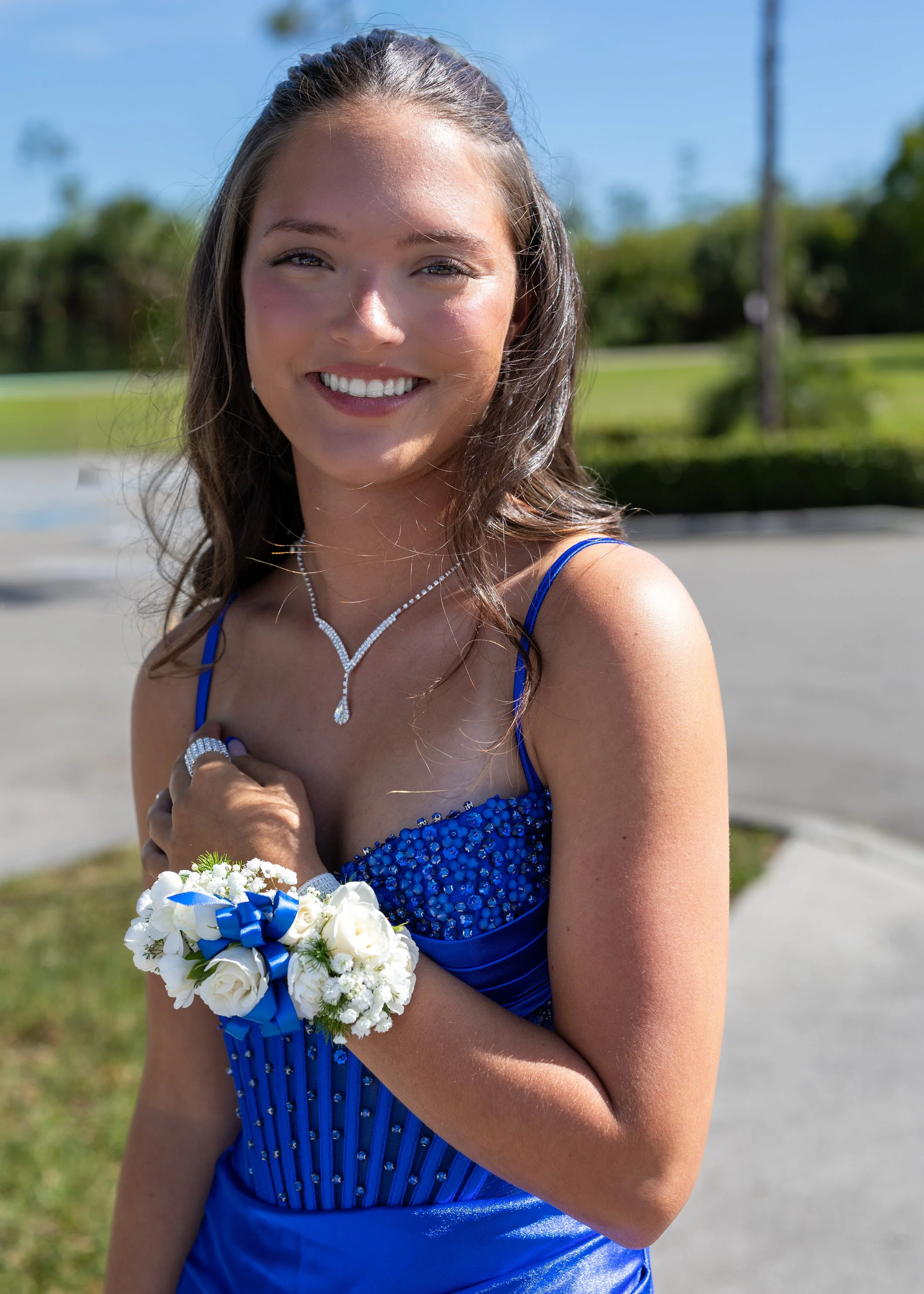 A young woman in a blue dress holding a bouquet of white flowers with blue ribbons, outdoors on a sunny day, smiling at the camera.