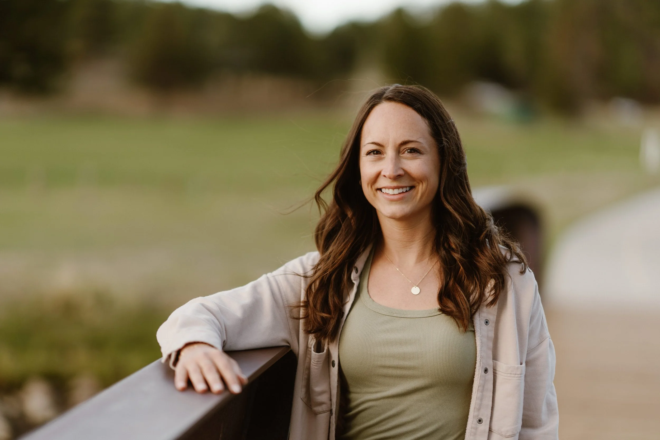 A smiling woman with long wavy brown hair is sitting on a park bench, wearing a light-colored jacket over a green top, with trees and a grassy area in the background.
