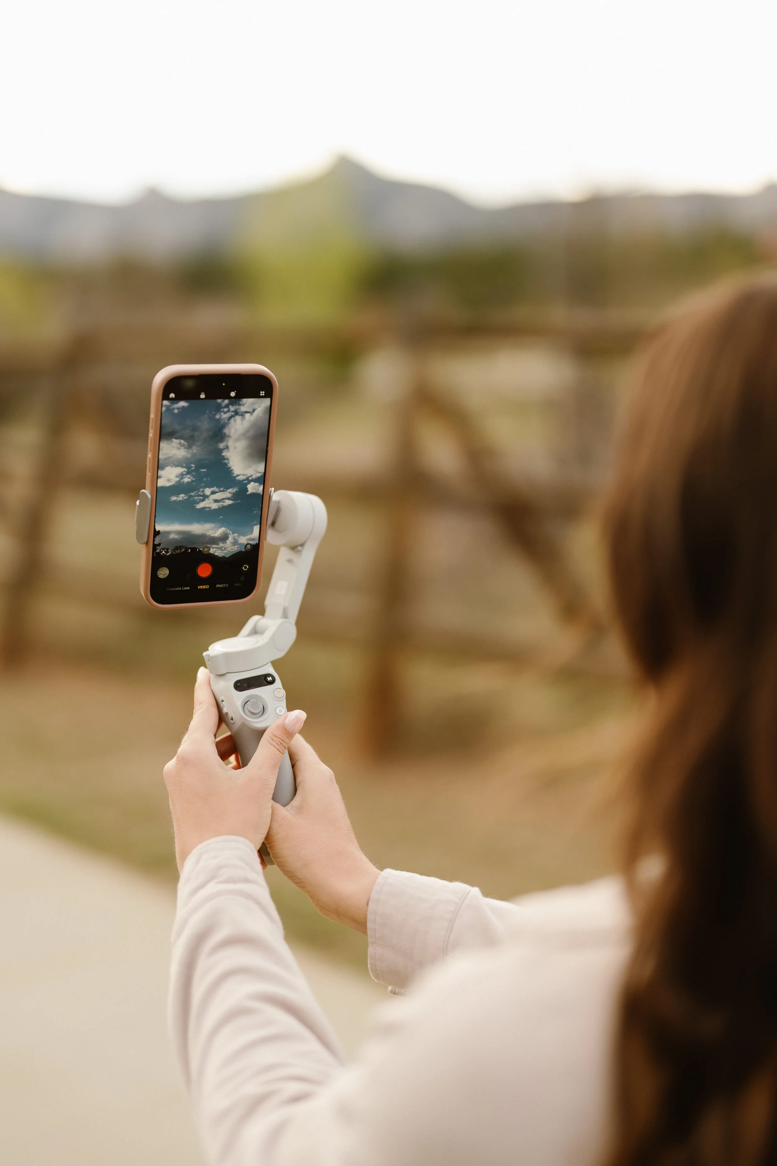 A person holding a smartphone on a stabilizer, taking a photo or video of a cloudy sky outdoors.