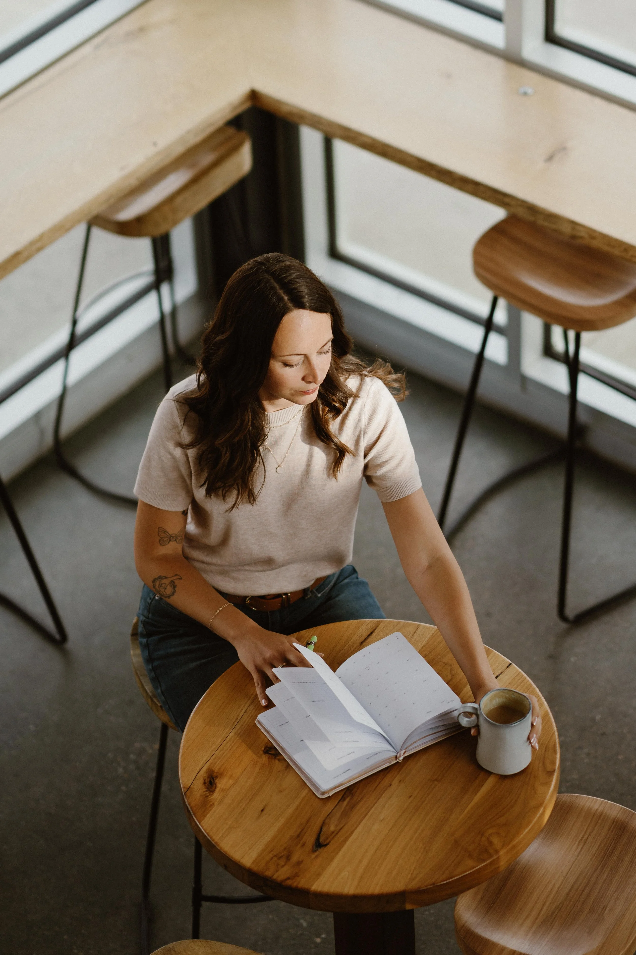 A woman with dark brown hair and tattoos on her left arm sitting at a round wooden table reading a notebook and holding a pen. There is a mug of coffee and a book on the table. The setting appears to be a cozy cafe or cafe-style workspace with large windows and barstool seating.