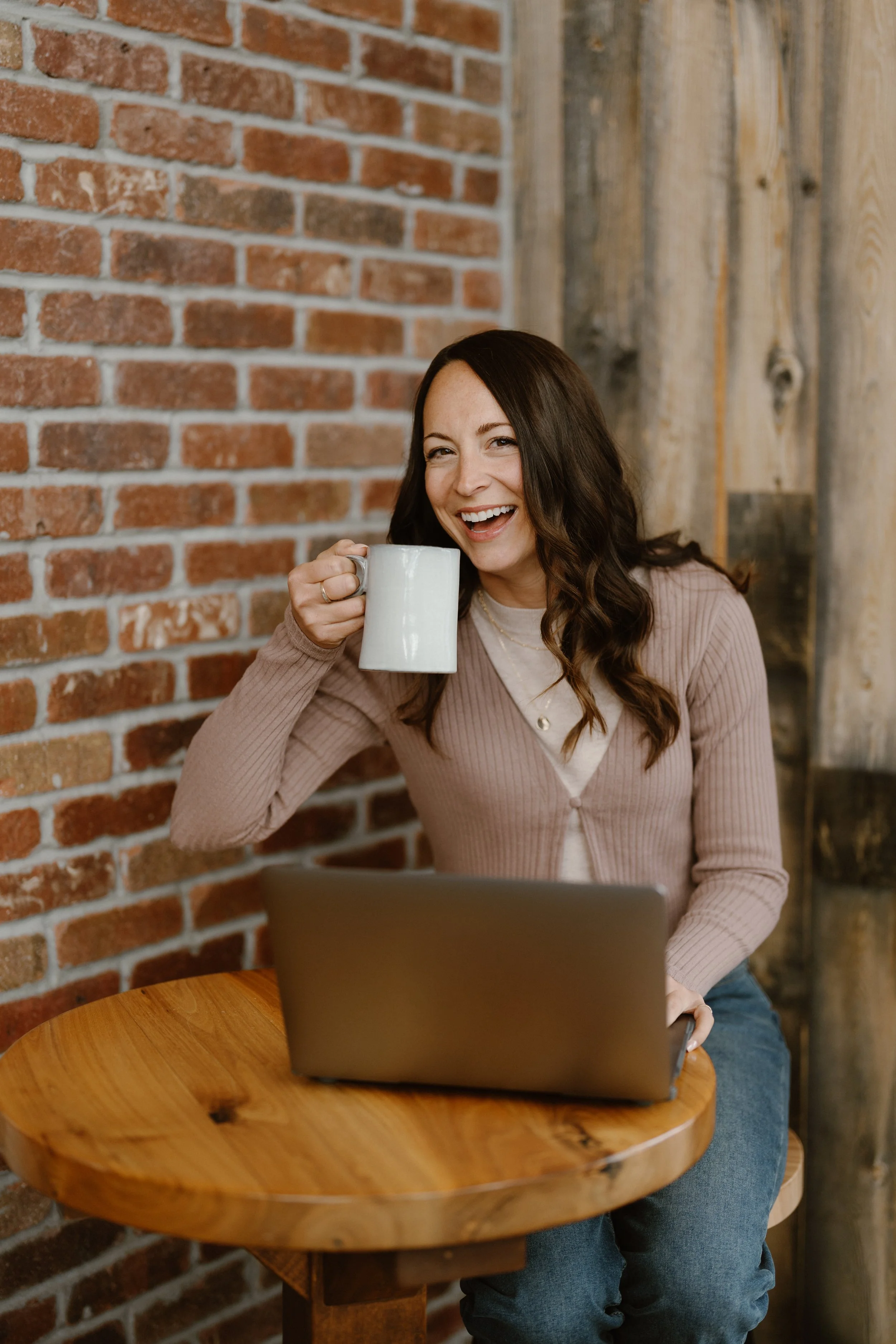 A woman sitting at a wooden table with a laptop, smiling and holding a mug in a cozy cafe with brick and wooden walls.