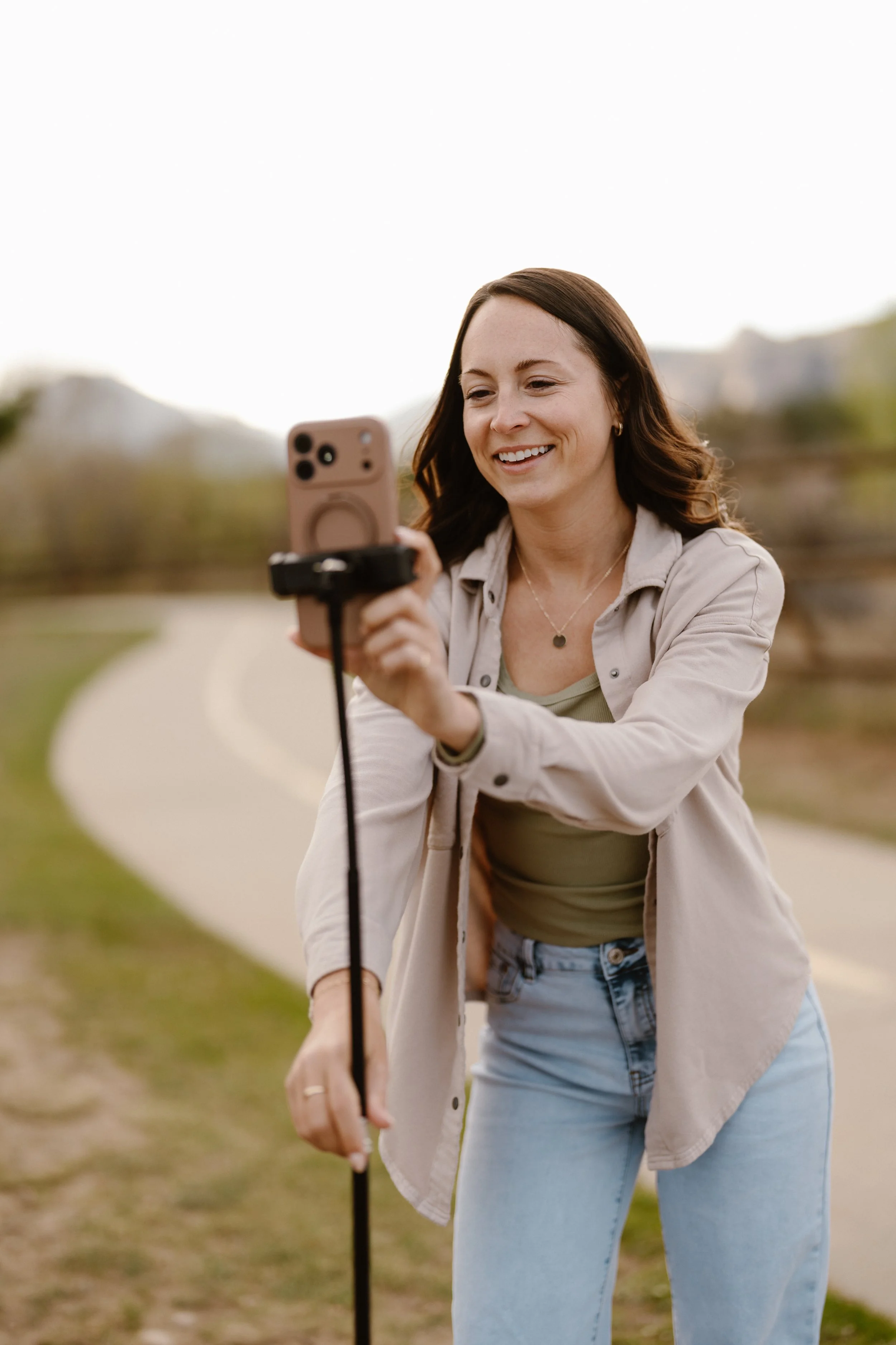 A woman with brown hair and light skin smiling while taking a photo with her smartphone outdoors on a pathway, with blurred greenery and trees in the background.