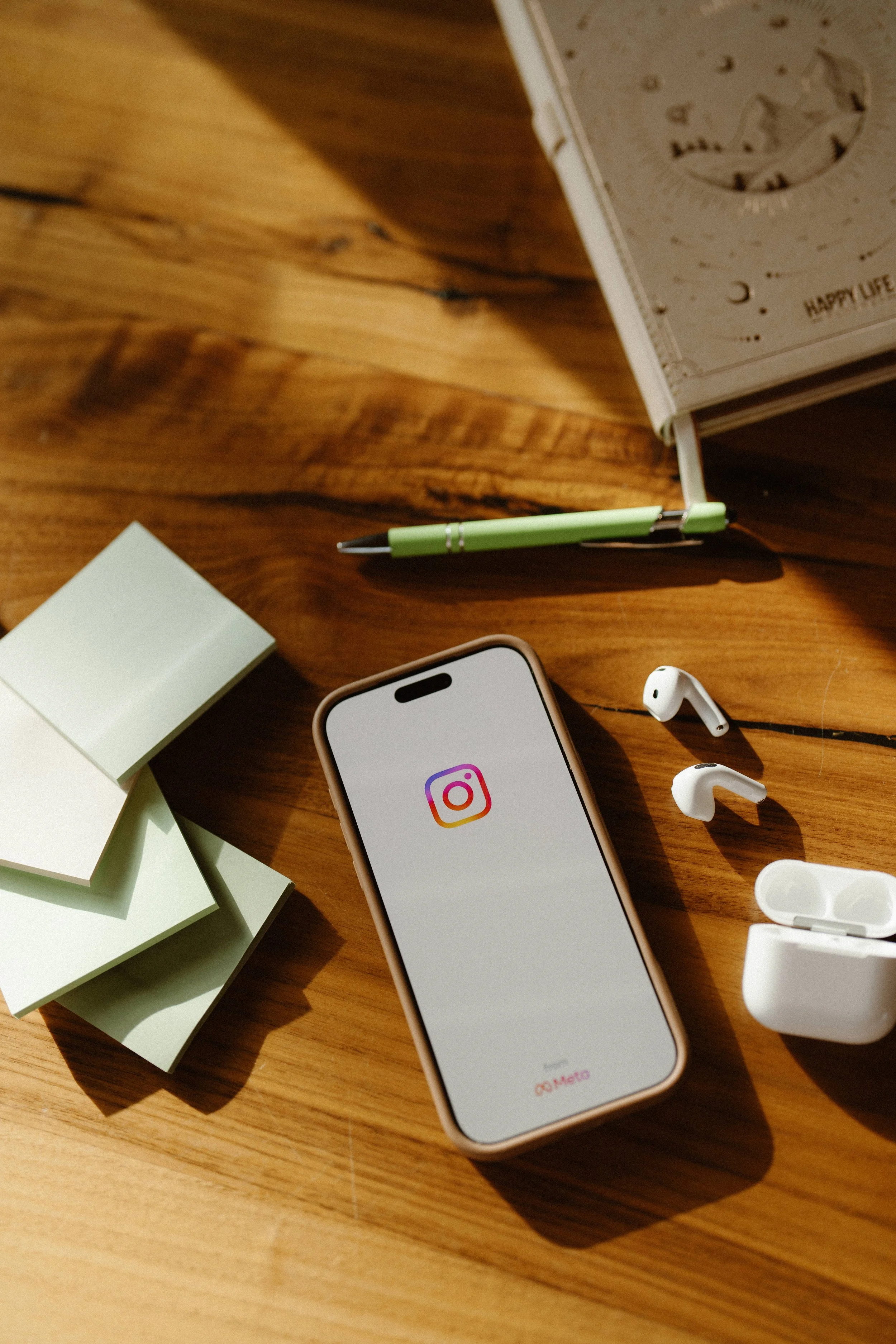 A wooden desk with a smartphone displaying the Instagram logo, a green pen, a partially visible white earbud, a container of white earbud tips, and a book with a celestial illustration.