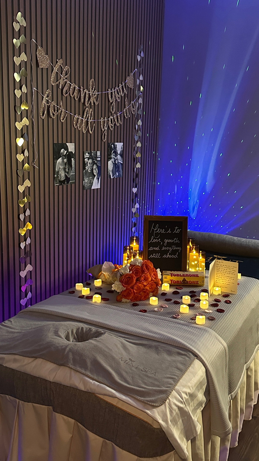 A decorated bed at a wedding or romantic celebration, with a bouquet of orange roses, candles, rose petals, and a framed chalkboard with a handwritten message, against a wall with fairy lights, photos, and a 'Thank you for all you do' banner.