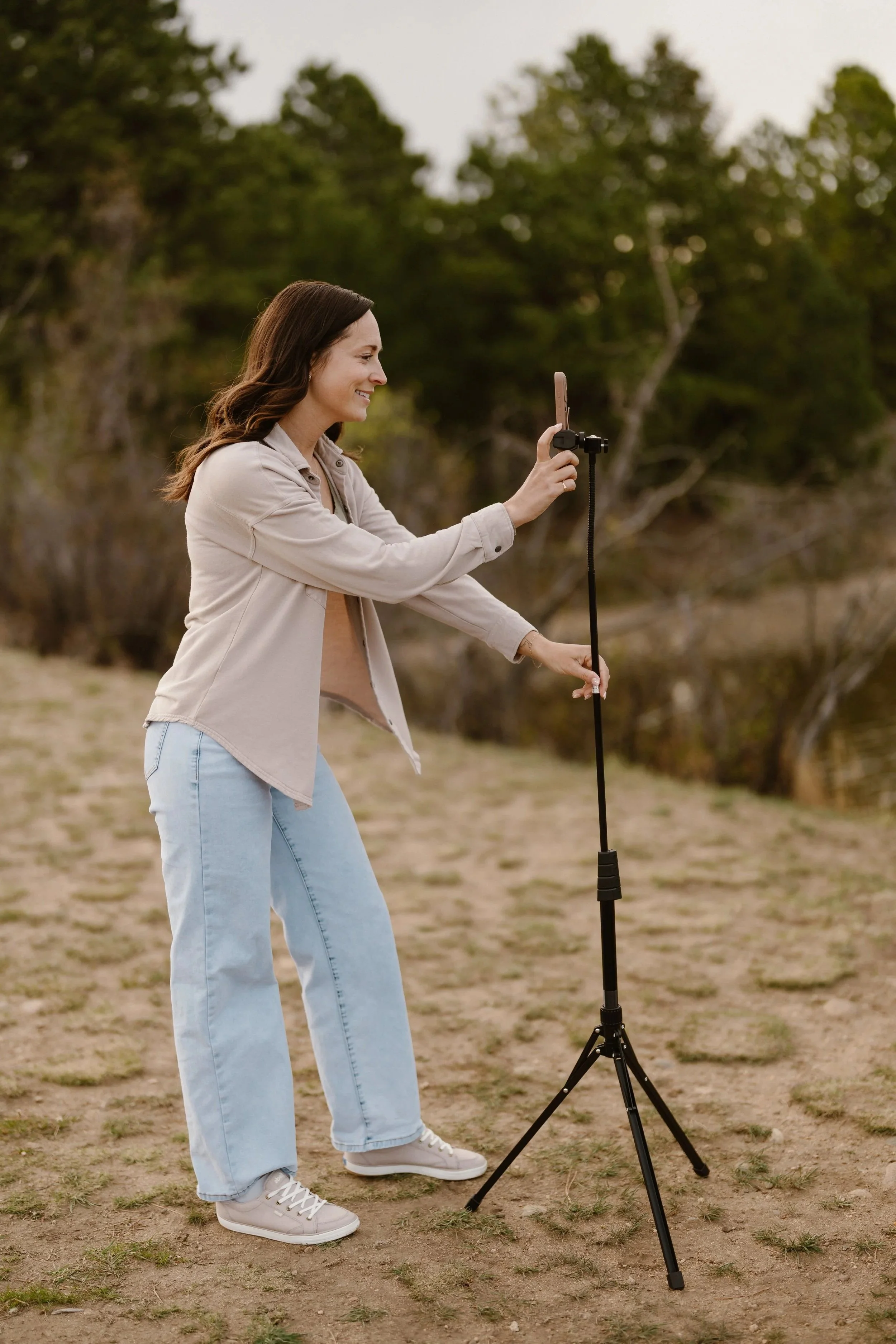 Woman with brown hair in beige jacket and light blue jeans standing outdoors on a dirt patch, smiling, and holding a smartphone on a selfie stick.