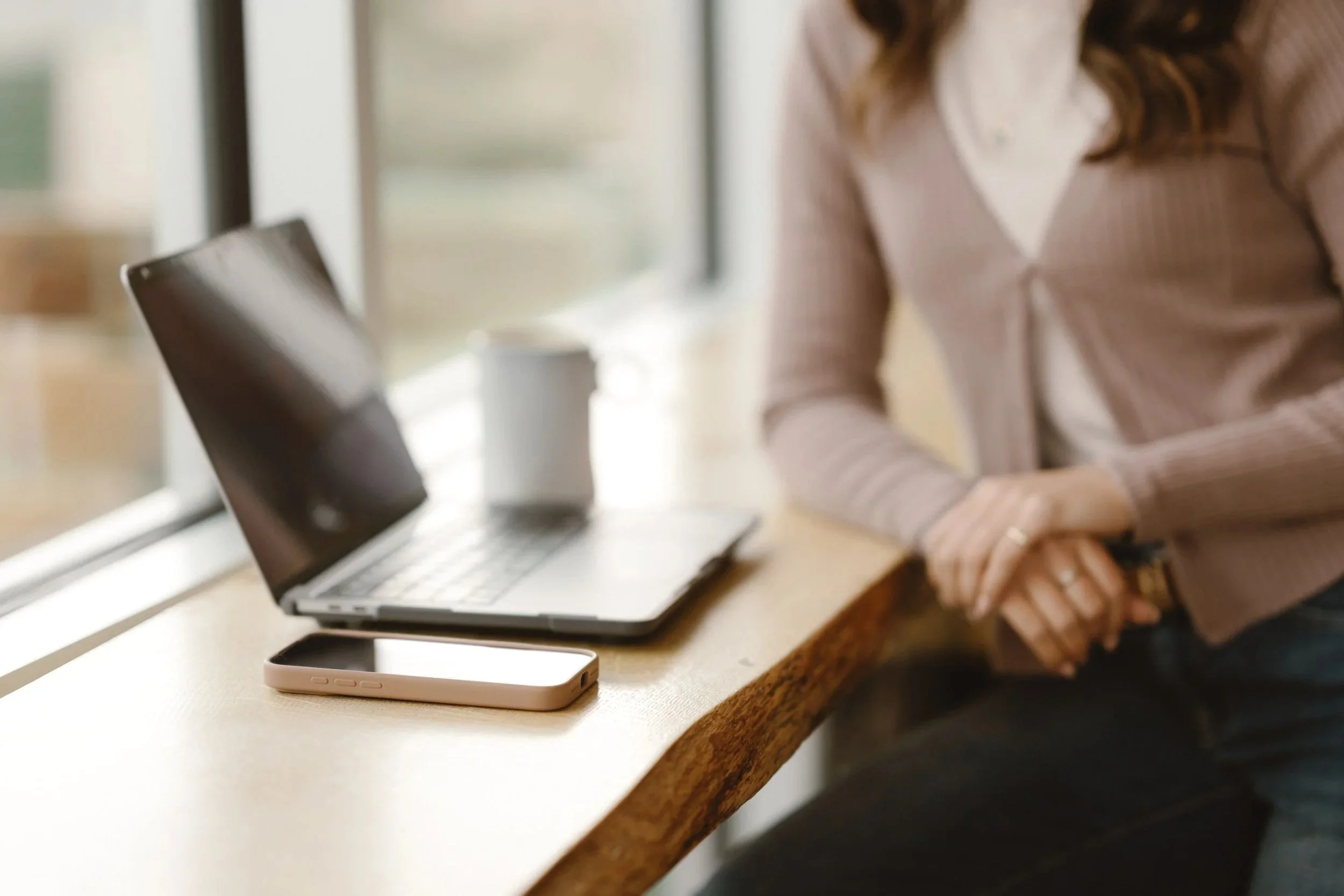 A social media manger sitting at a wooden table with a laptop and a smartphone, near a window working on content creation.