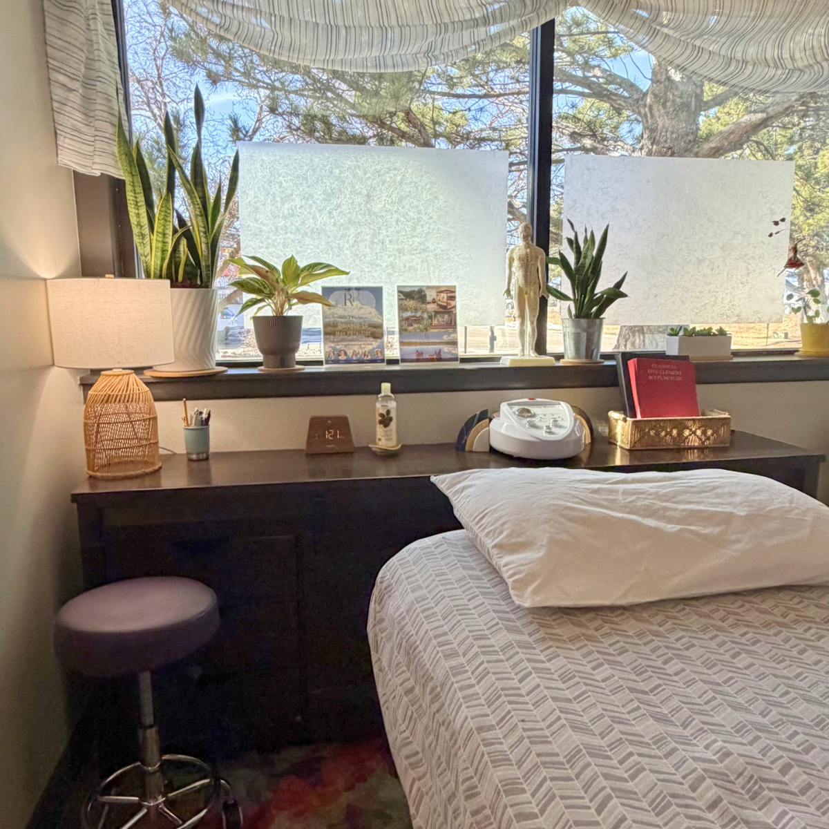 A bedroom with a bed, a dark wooden desk, and a window with frosted glass. The desk has potted plants, books, a lamp, a clock, a bottle of hand sanitizer, and a massage device. There is a purple stool next to the desk. The window has some frosted areas and tree branches visible outside.