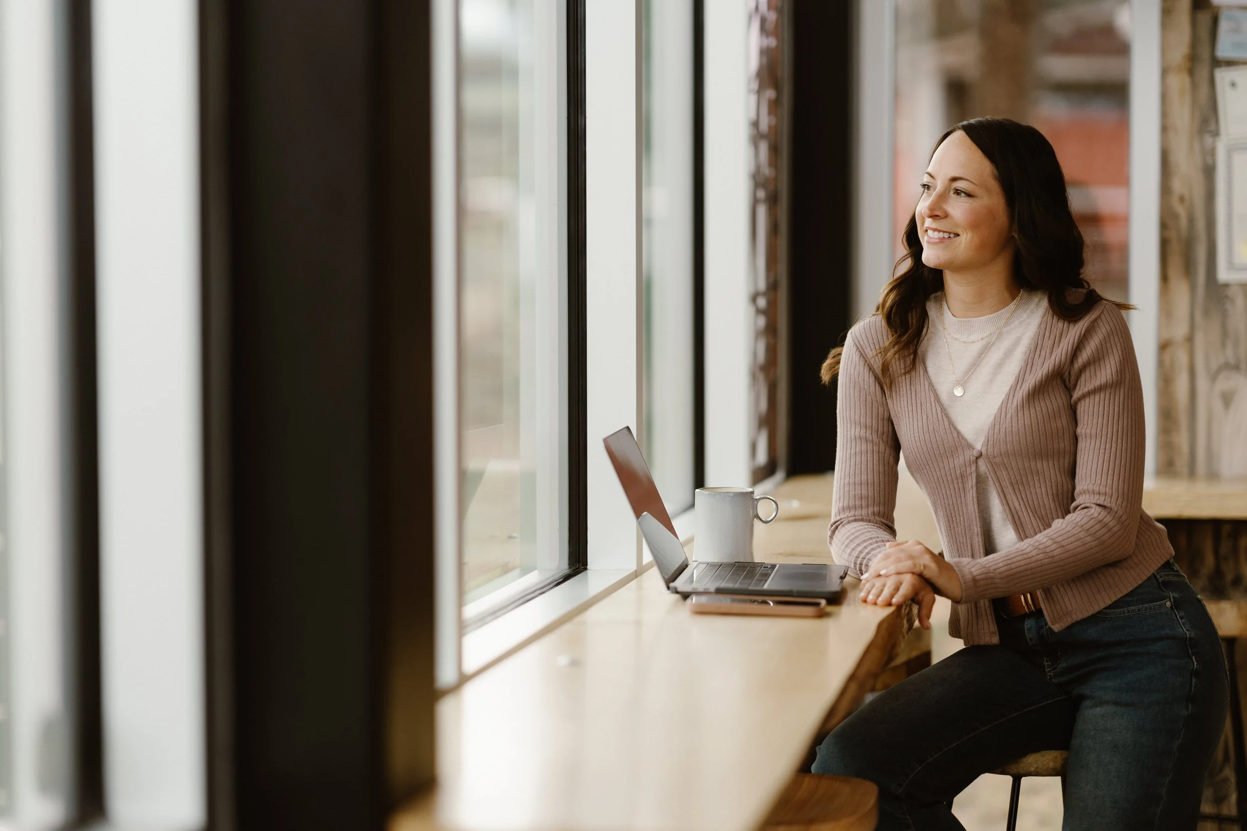 A woman with long dark hair smiling and working on a laptop at a counter near a window in a cafe.