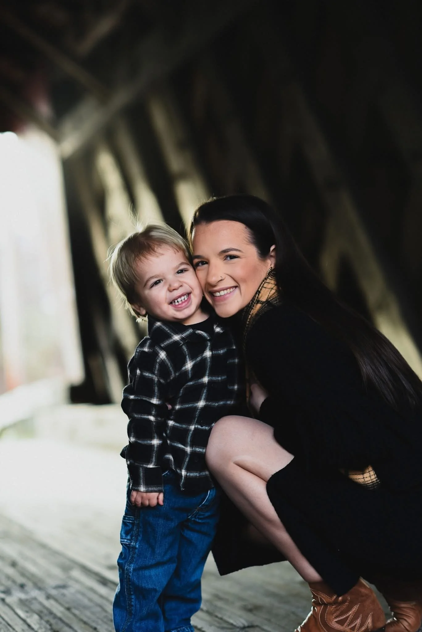 A woman and a young boy smiling and hugging inside a barn with wooden beams.