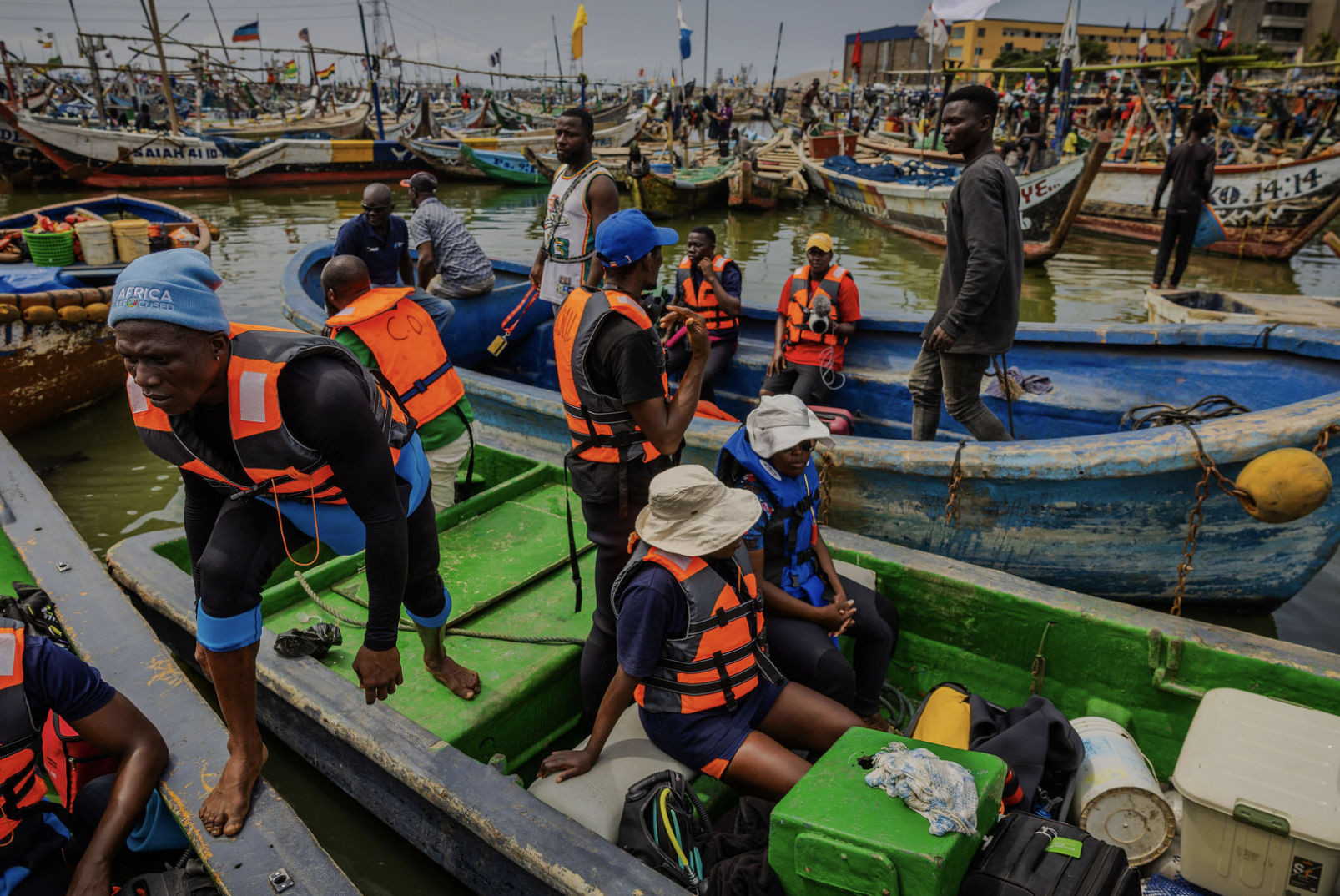 People are preparing boats at a busy harbor, with numerous fishing boats docked in the background, some wearing life jackets and hats.
