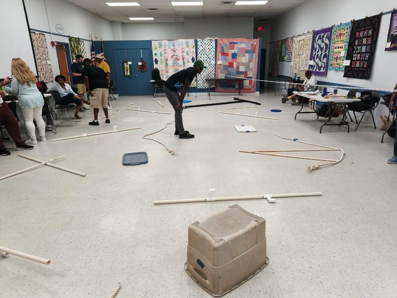 People in a room with handmade quilts on the walls and objects like pipes, ropes, and boxes arranged on the floor, suggesting a group activity or workshop.