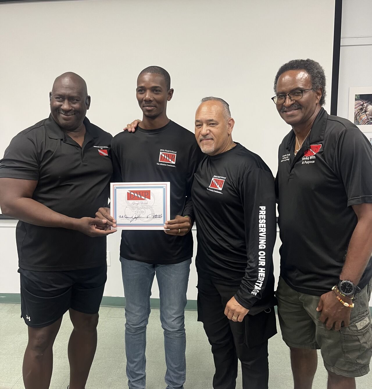 Four men standing together, one holding a certificate. All four are wearing black polo shirts with logos, smiling, in an indoor setting.