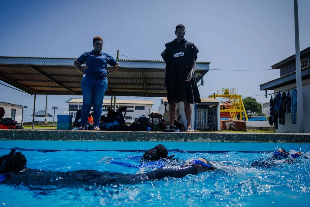 Swimmers practicing in an outdoor swimming pool with two women and one man standing on the poolside observing.