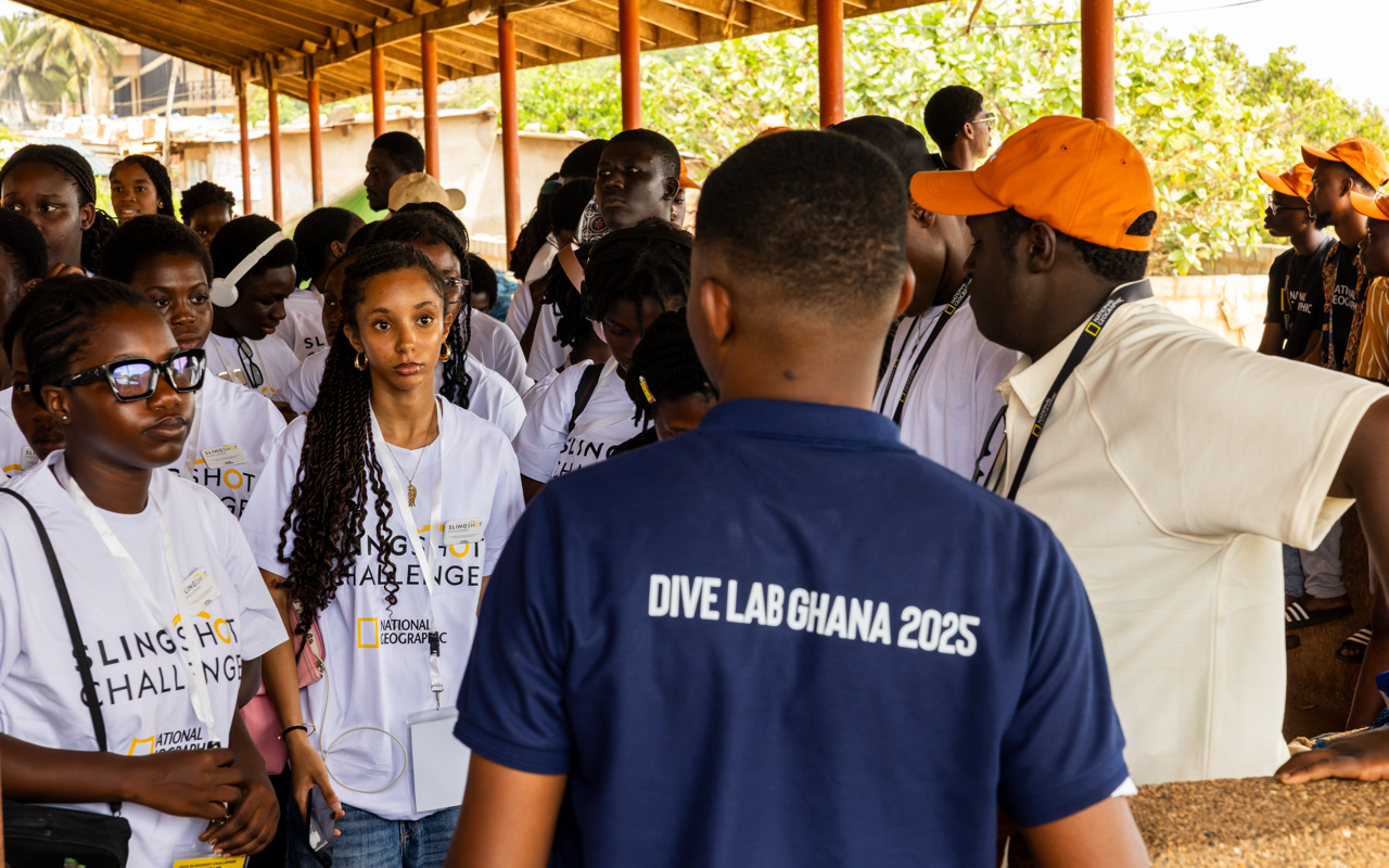 Group of young people wearing white T-shirts with 'SLINGSHOT CHALLENGE' and 'NATIONAL GEOGRAPHIC' logos, standing outdoors under a roof, listening to a man in a white shirt and a person in a dark blue shirt with 'DIVE LAB GHANA 2025' on the back, during an educational event.