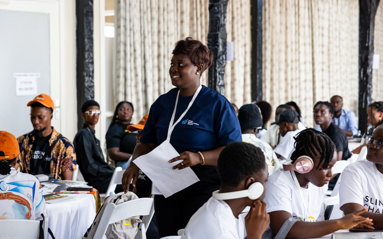 A woman standing in a conference room filled with seated attendees, some of whom are wearing headphones; she holds papers and wears a navy blue shirt with a logo.