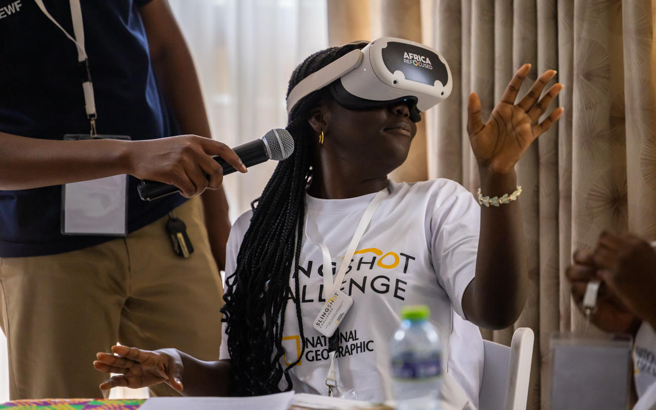 A woman with braided hair wearing a virtual reality headset labeled 'Africa ReFocused' and a white T-shirt, raising her hand while another person holds a microphone near her.