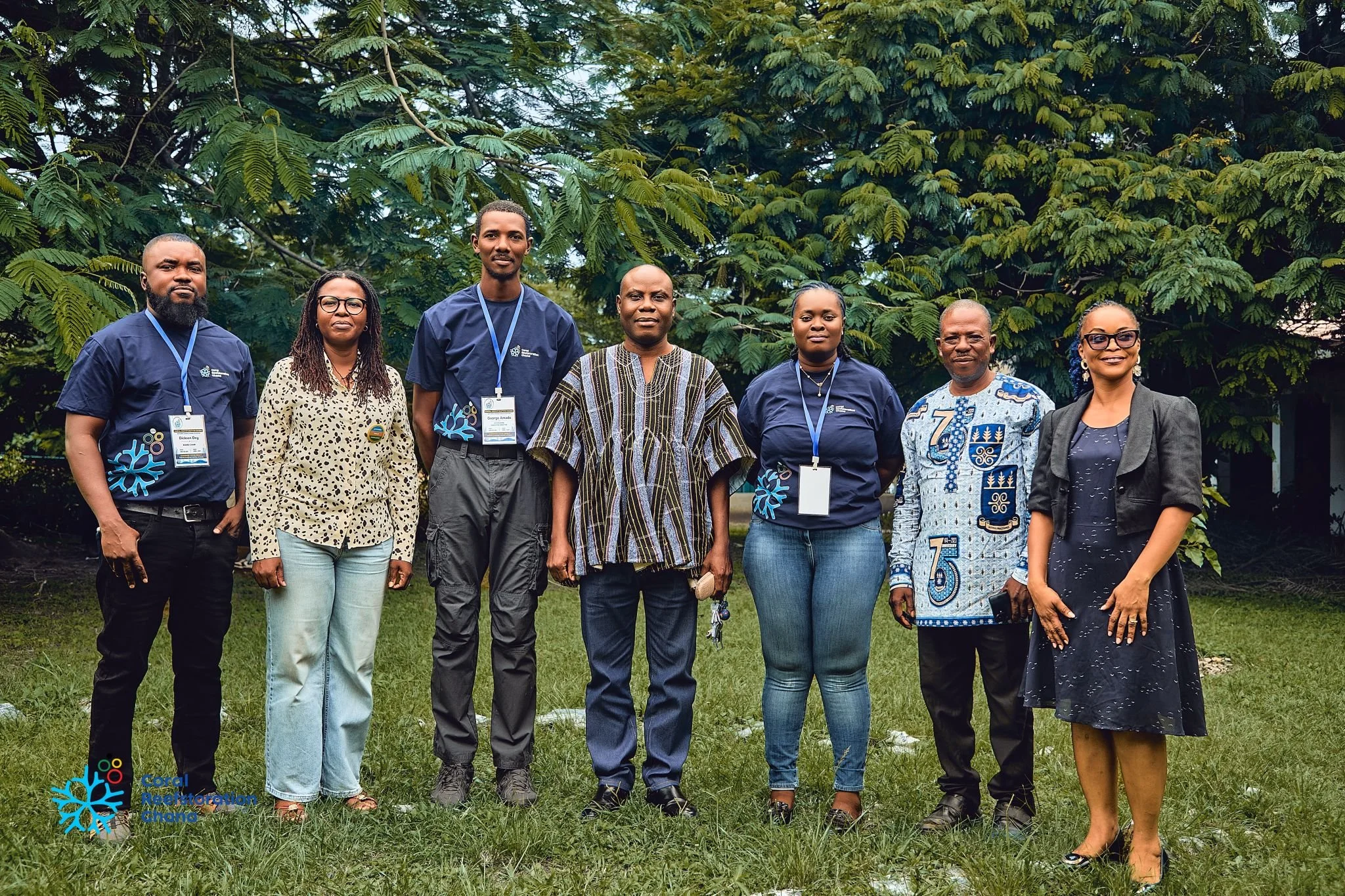 Group of seven people standing outdoors in front of green foliage, some wearing conference badges and smiling.