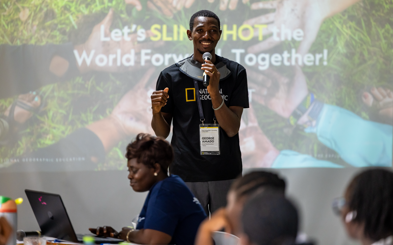 A young man with a microphone smiling during a presentation at an event with a large screen behind him showing a message about taking action together for the world. Several people are seated at tables in front of him, one woman typing on a laptop.