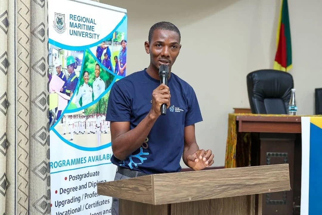 A man speaking at a podium during an event at the Regional Maritime University. In the background, there is a banner displaying the university's name and programs, along with a chair and flag.