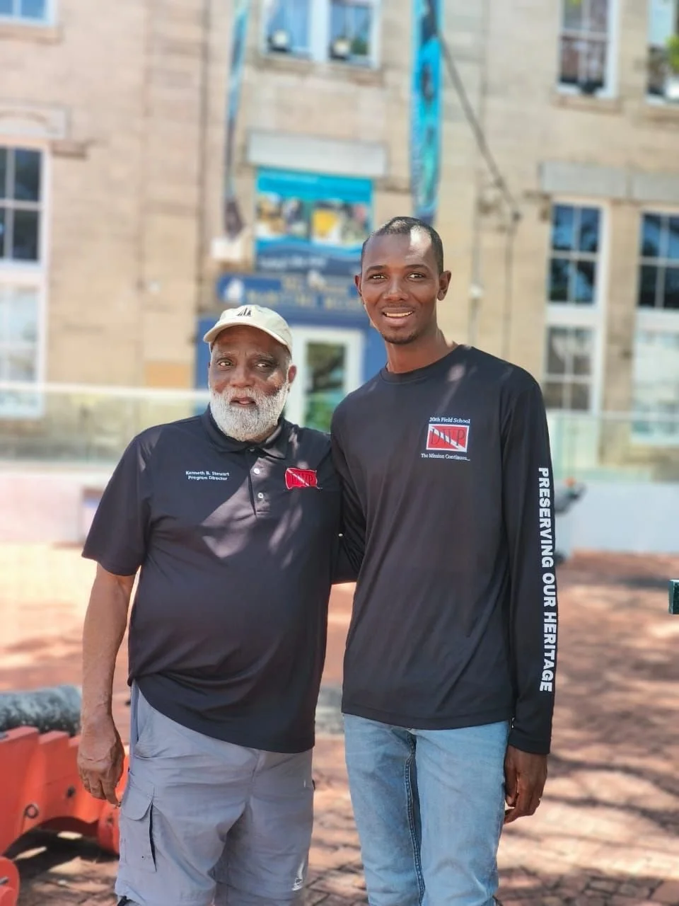 Two men standing outdoors in front of a building, smiling, wearing black shirts with logos. One has a white beard and cap, the other is younger with short hair. They appear to be part of a community or heritage preservation event.