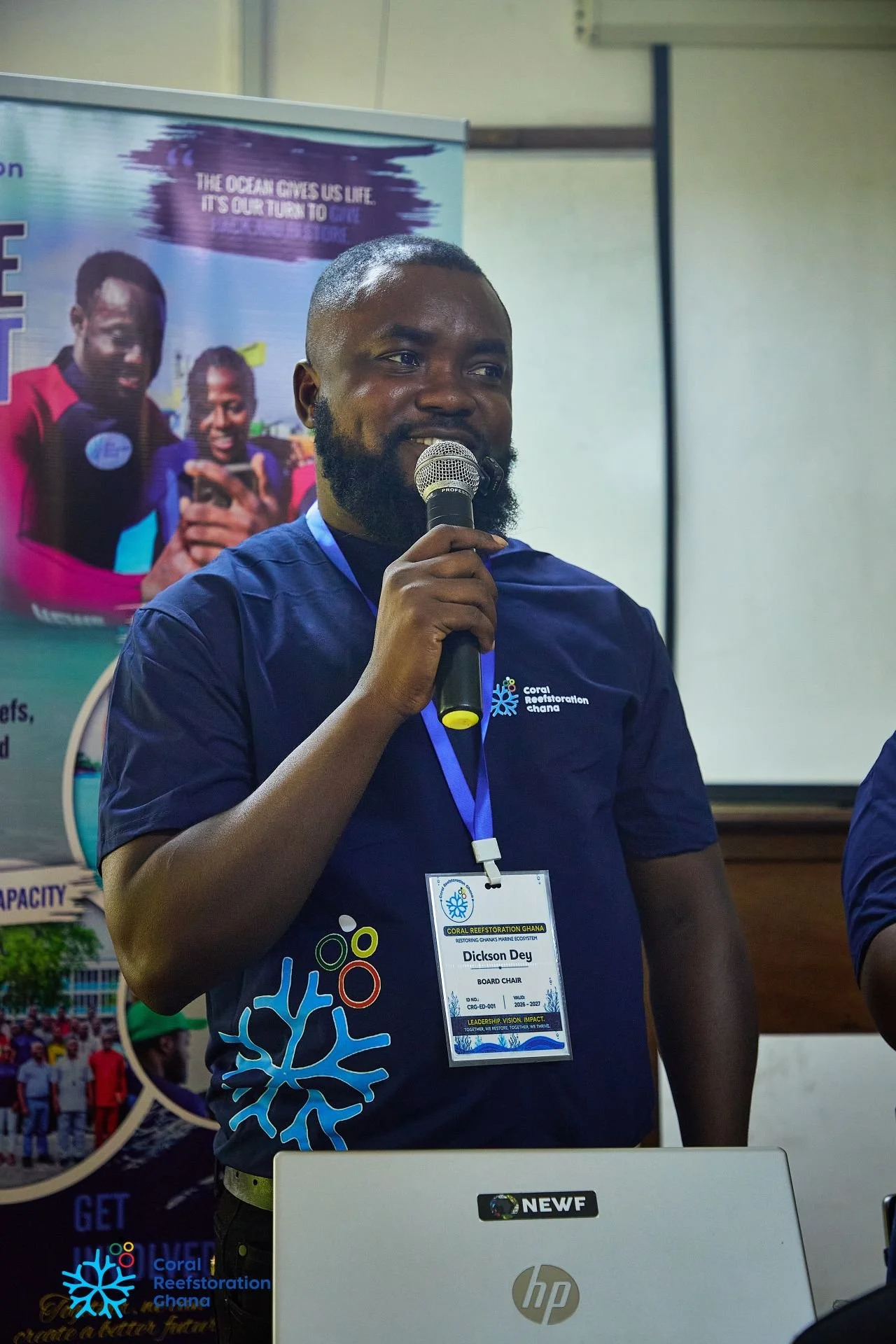 A man speaking into a microphone at a conference, wearing a dark blue shirt with a name tag that reads 'Dickson Dey'. Behind him is a colorful banner with people taking photos and text promoting corporate reef restoration in Ghana.