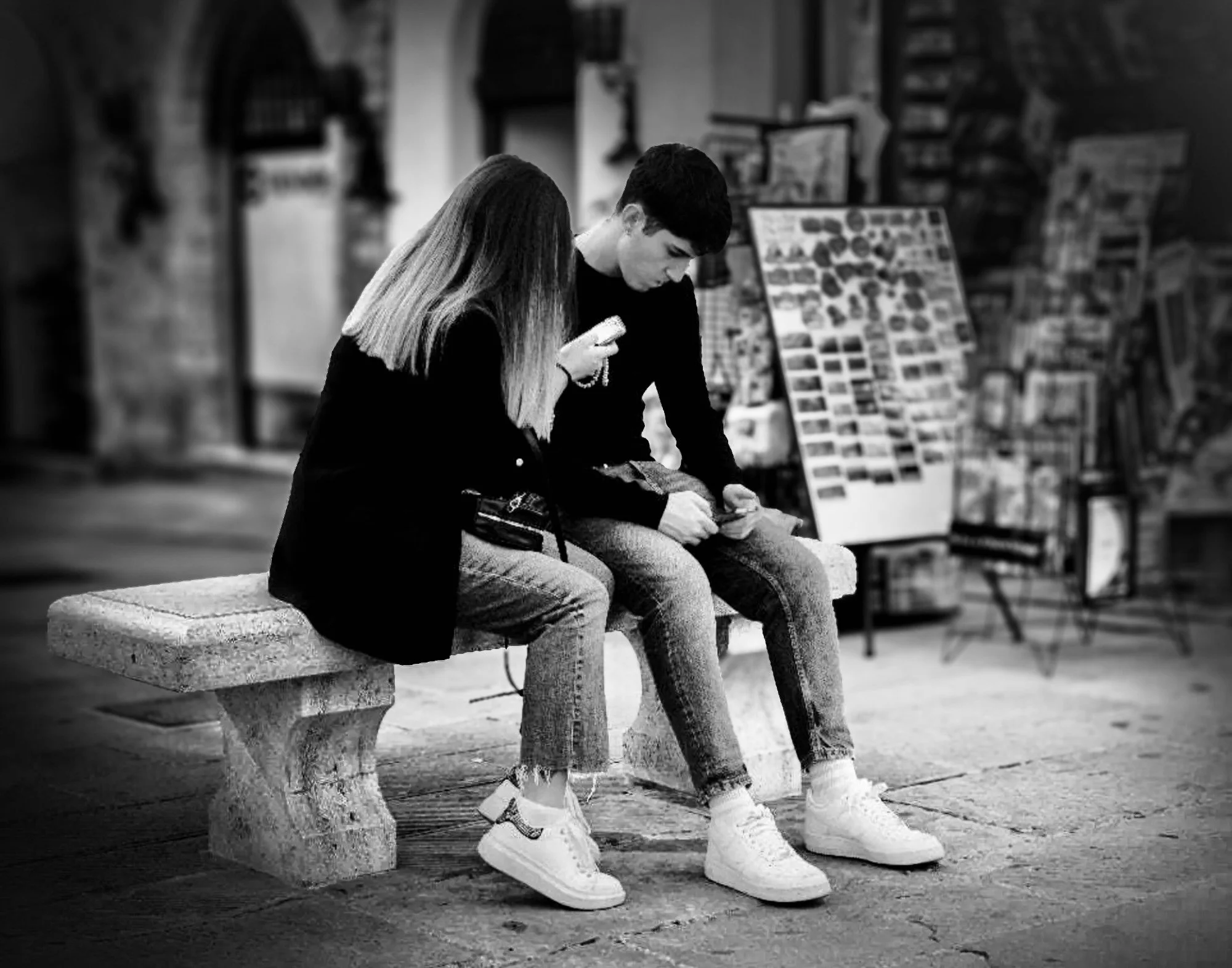 A young man and woman sitting on a stone bench in an Italian piazza looking at their smartphones in the waning afternoon light.
