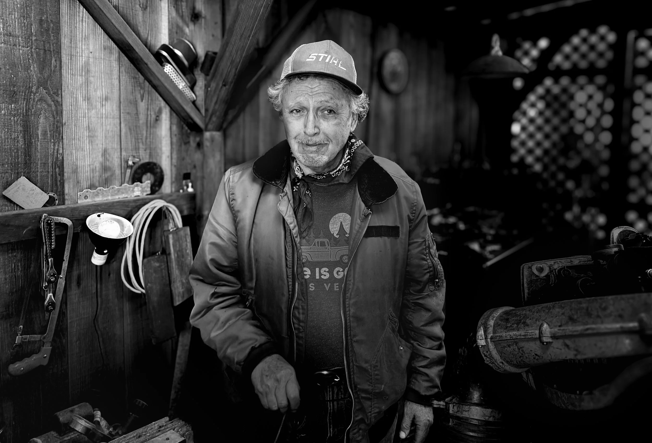 Black and white photo by John Patrick Weiss of his father-in-law, and older man wearing a winter jacket and baseball cap and standing inside his workshop.