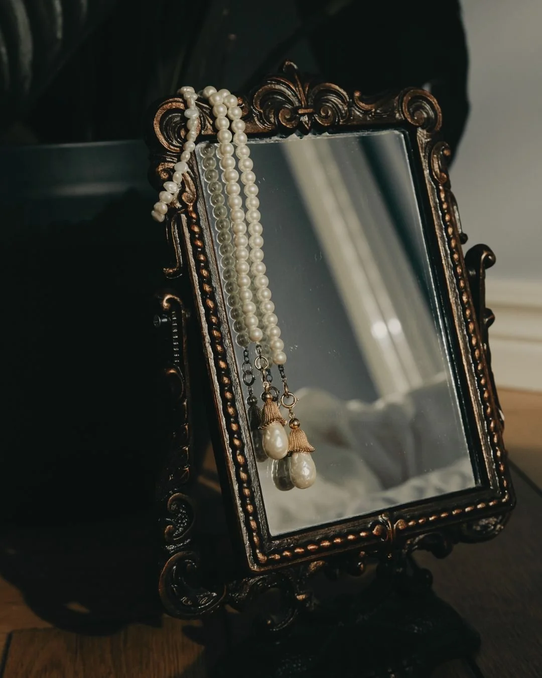 Pearl necklace and pearl earrings hanging on an ornate, vintage mirror with a dark wooden frame.