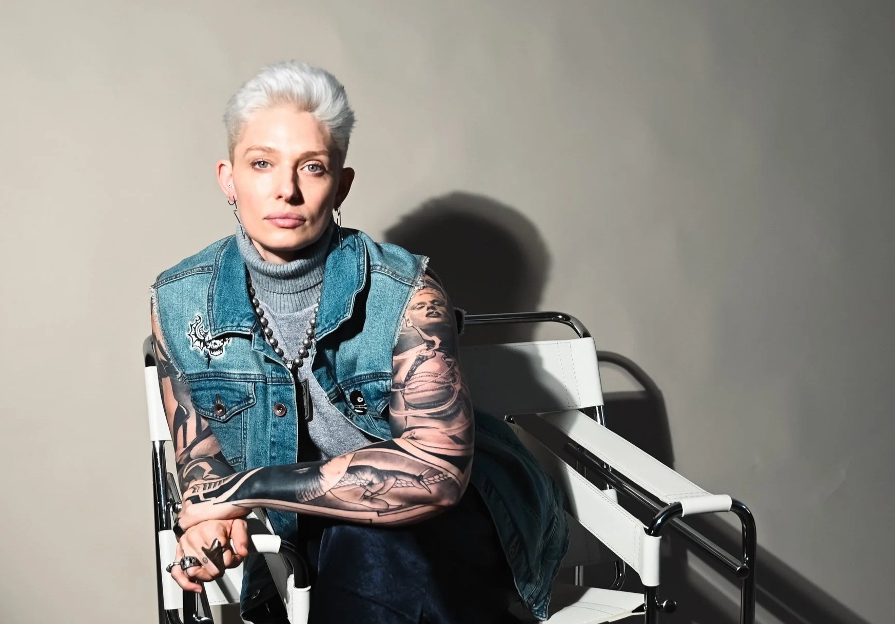 A person with platinum blonde hair, tattoos on both arms, wearing a denim vest over a gray turtleneck, and jewelry, sitting with crossed arms on a wheelchair against a plain background with a shadow cast on the wall.