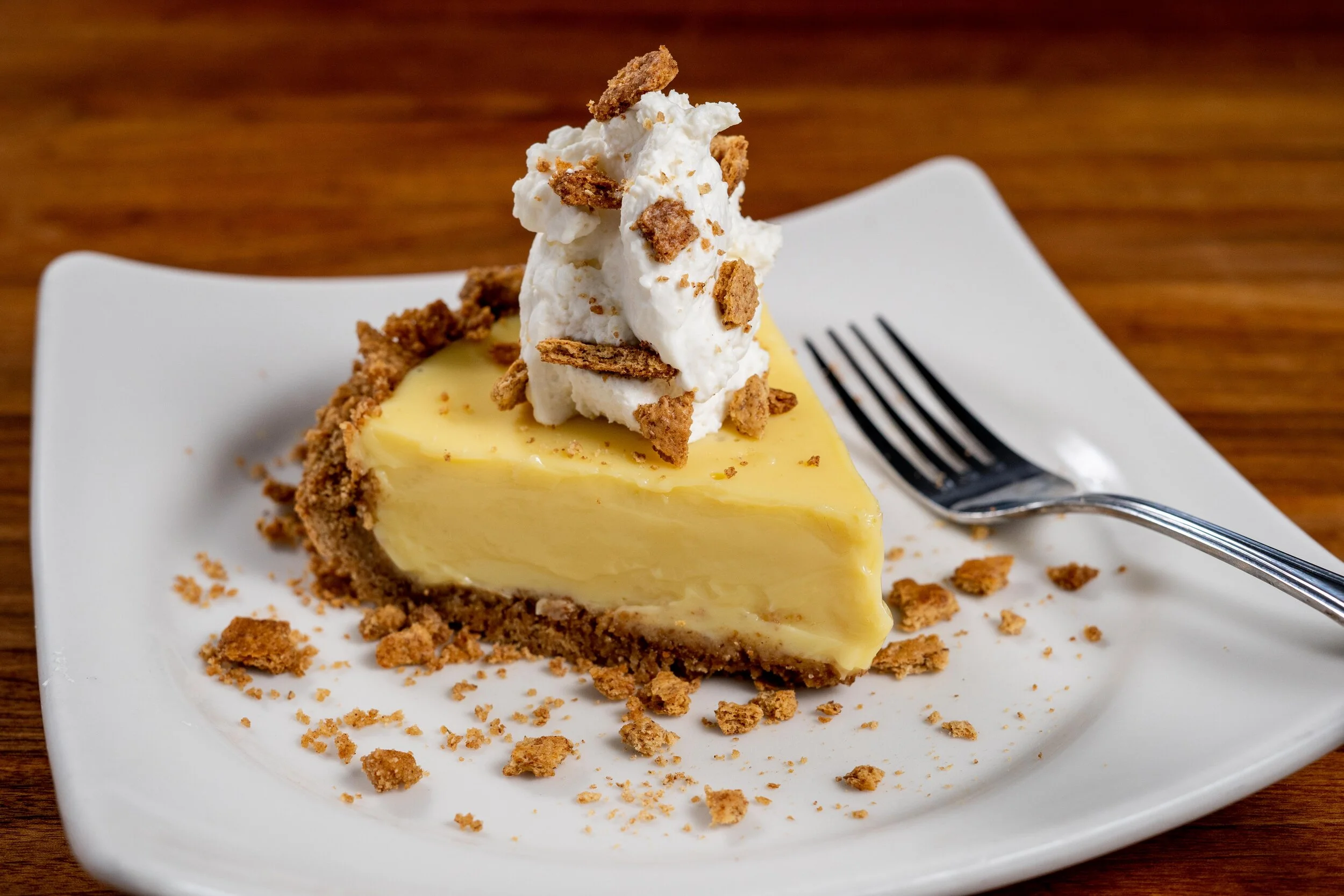 A slice of creamy cheesecake topped with whipped cream and crumbled cookies on a white square plate with a fork, placed on a wooden table.