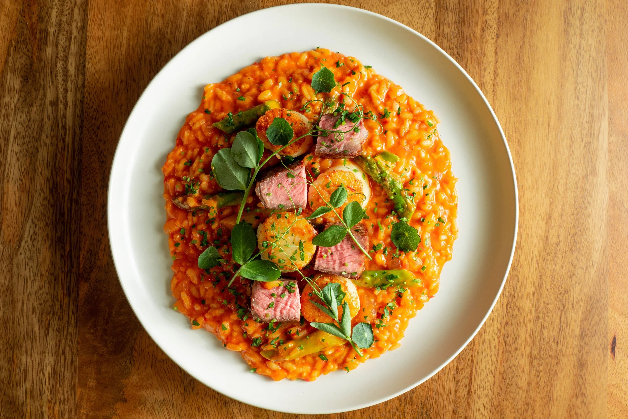 A white plate of risotto with vegetables and slices of beef, garnished with fresh herbs, on a wooden table.