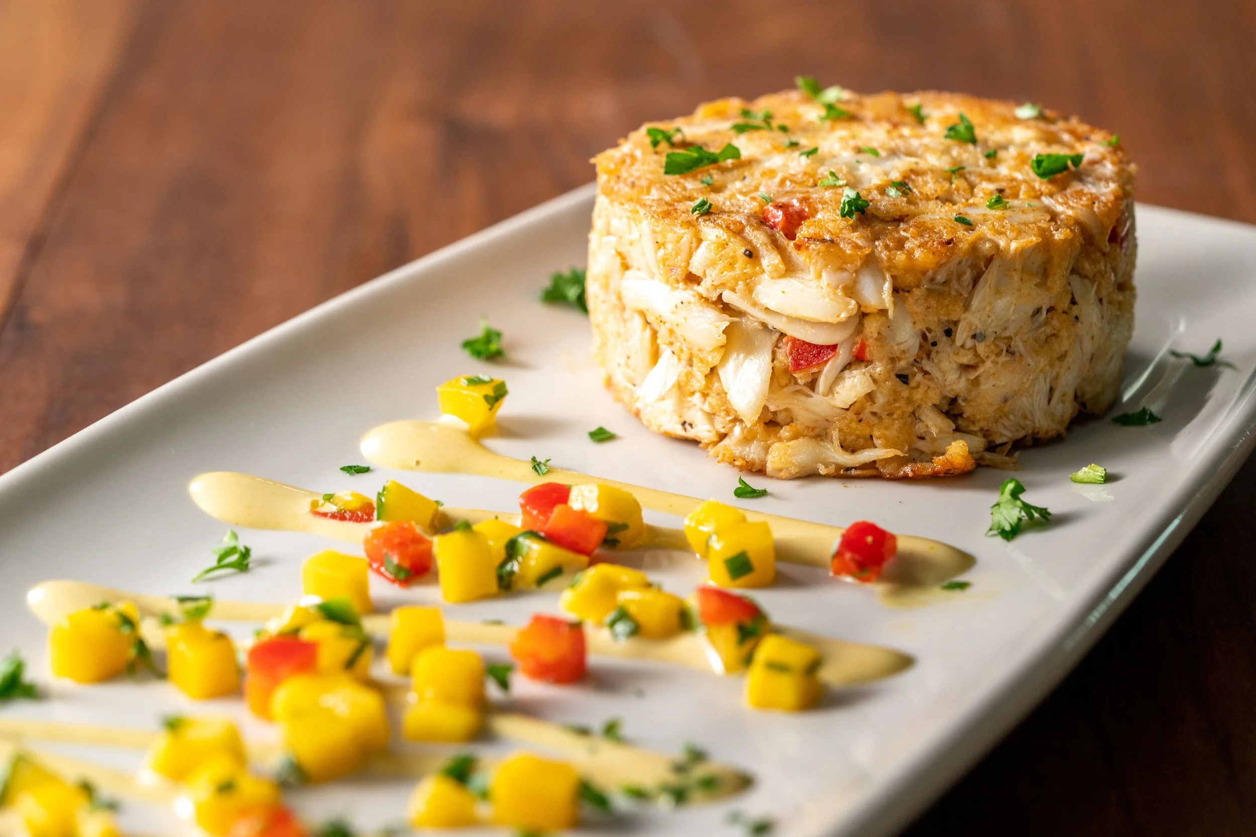 Crab cake on a white rectangular plate with mango and tomato salsa garnished with parsley and drizzled with a creamy sauce, placed on a wooden surface.