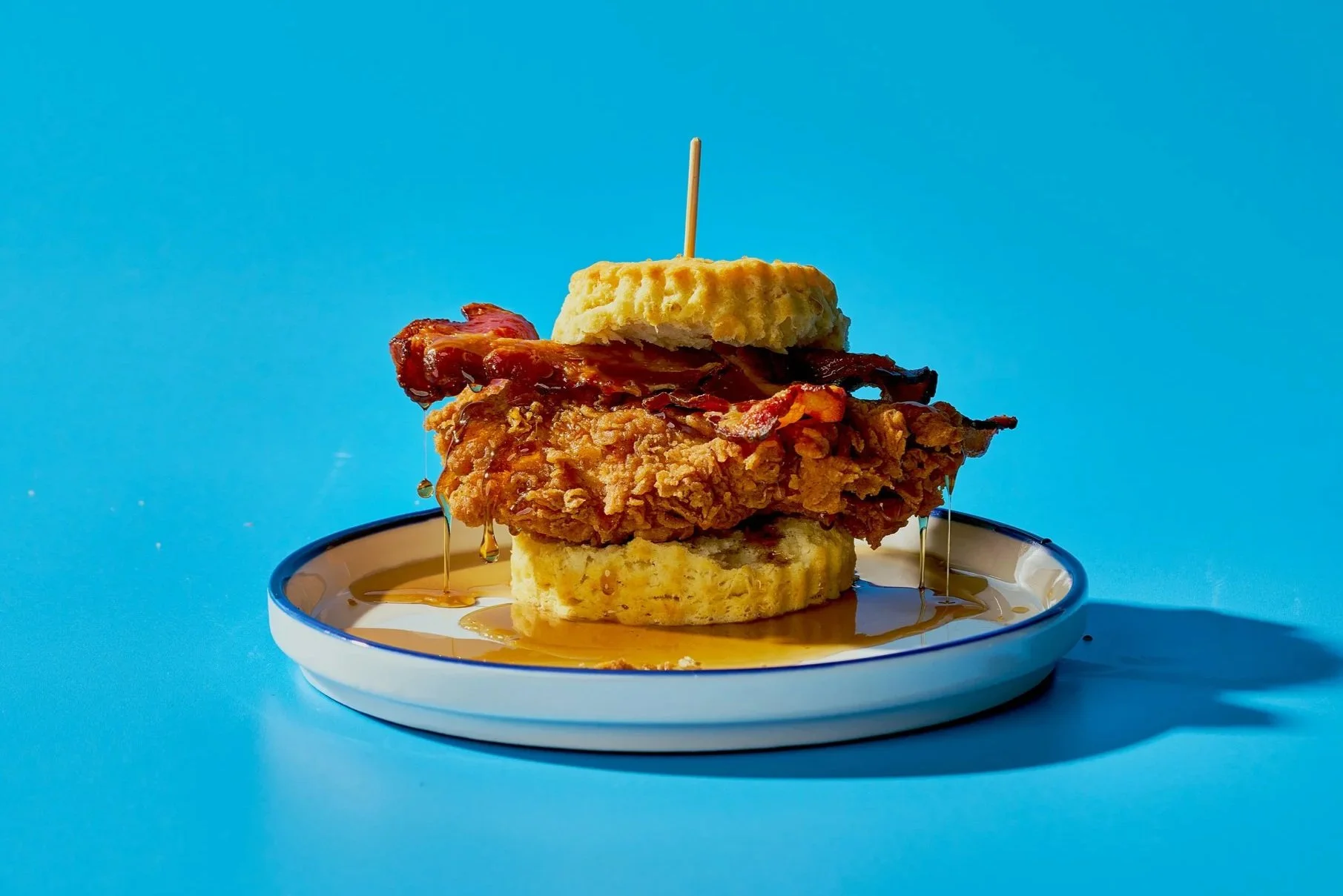 Fried chicken sandwich with maple syrup, bacon, and two waffle slices on a white plate against a blue background.