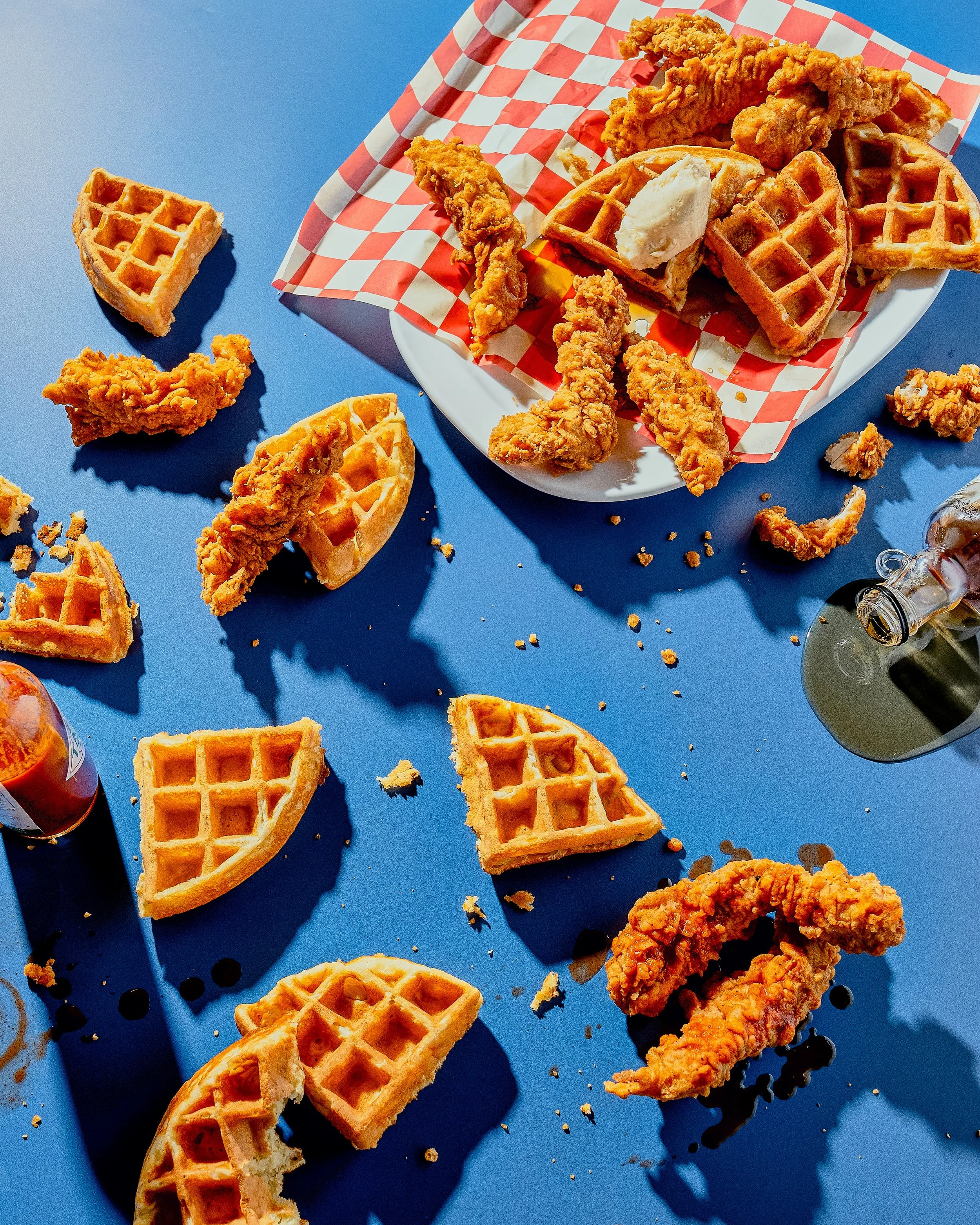 A plate of fried chicken tenders and waffles on a red and white checkered paper, with some waffles and chicken scattered on a blue surface.